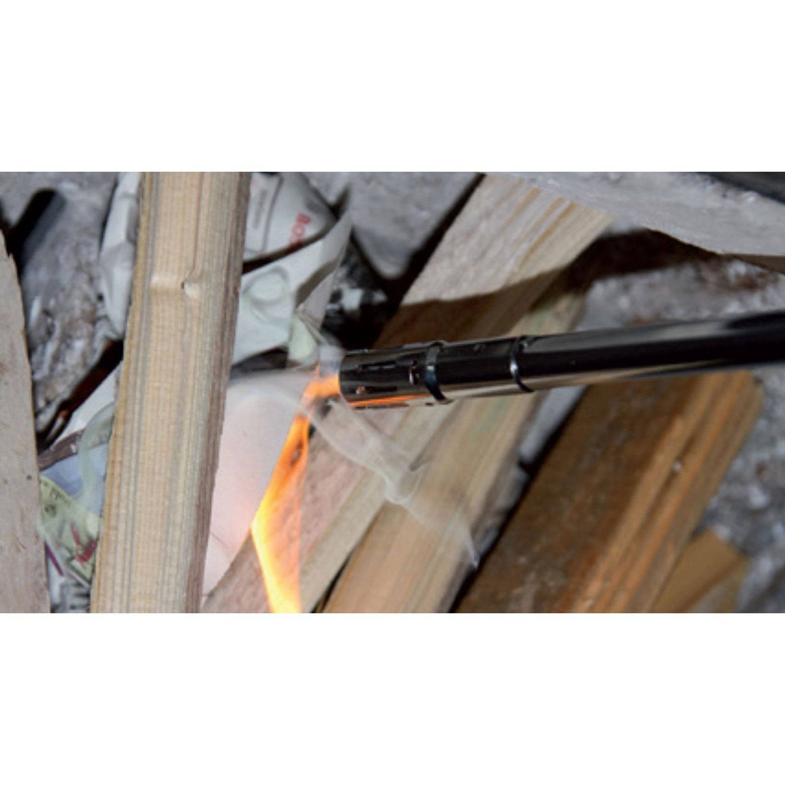 Nahaufnahme eines Metallfeueranzünders, der ein Feuer zwischen Holzscheiten und zerknittertem Zeitungspapier entzündet, wobei eine sichtbare Flamme zu sehen ist.