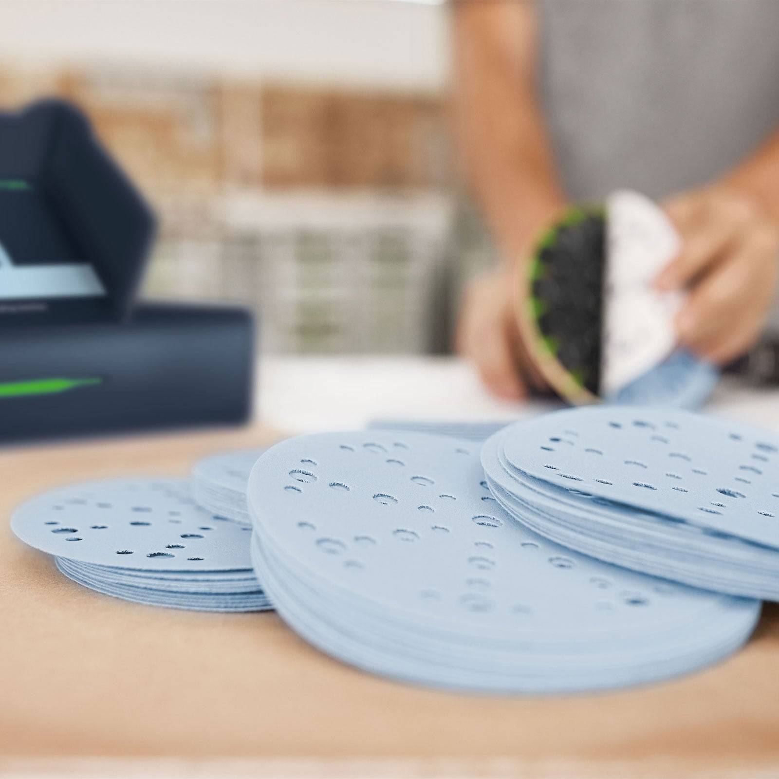 Ein Stapel blauer Schleifteller mit Löchern liegt auf einem Tisch. Eine Person hält einen Teller in der Nähe eines Schleifwerkzeugs.