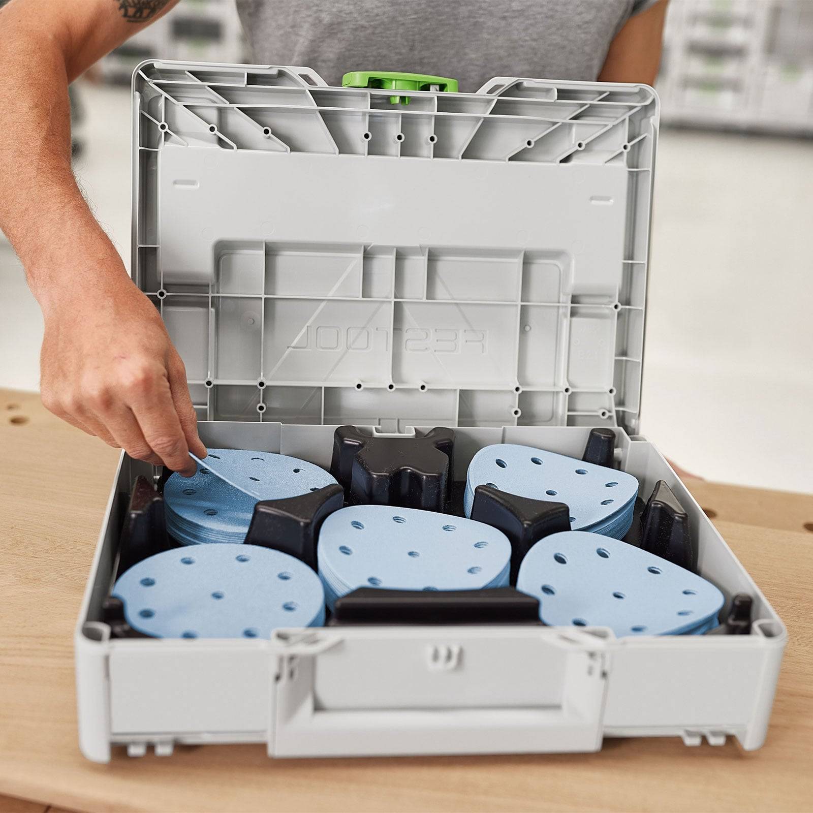 Eine Person ordnet blaue Schleifscheiben in einem offenen Werkzeugkoffer auf einem Holztisch an.