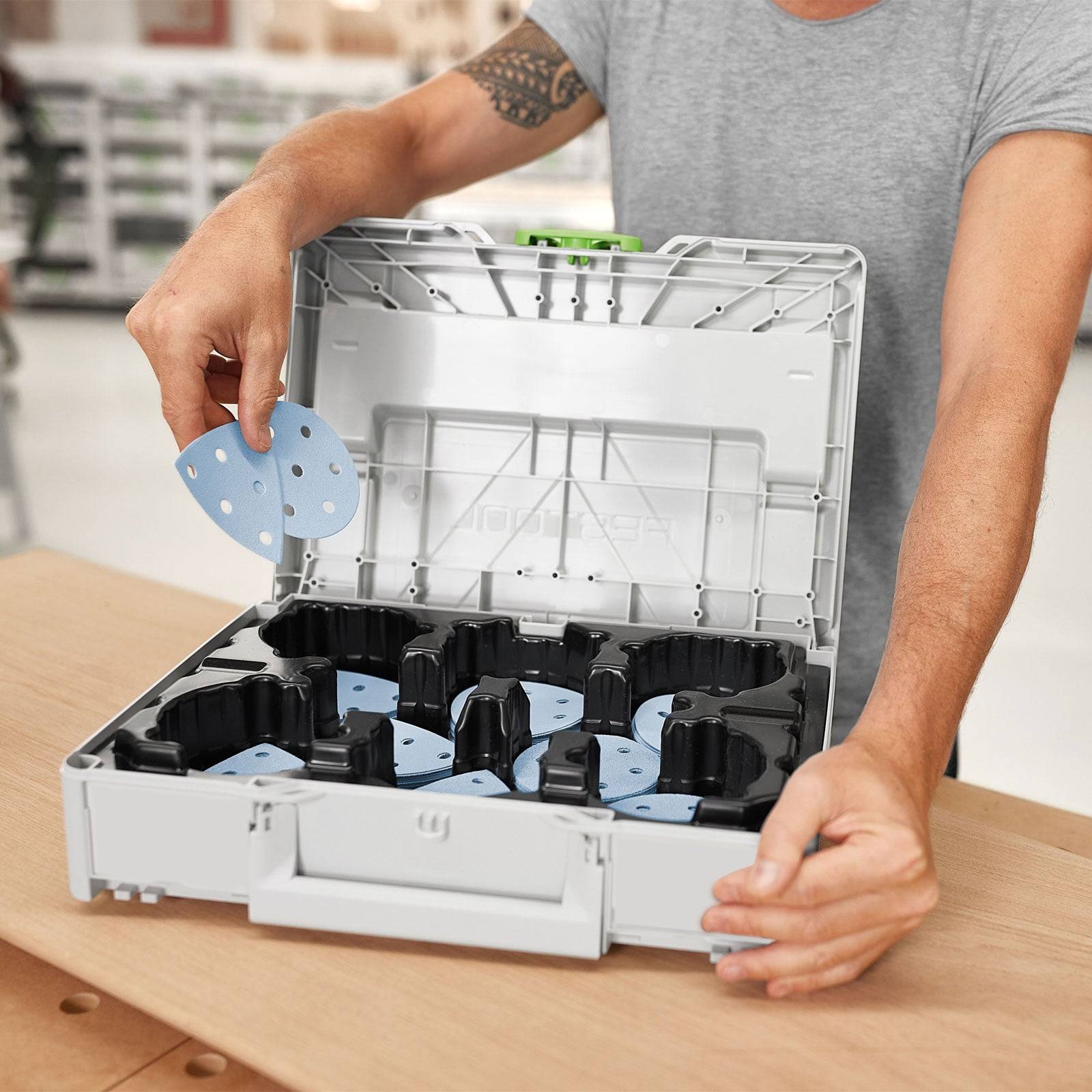 Eine Person legt eine runde blaue Schleifpapierscheibe in einen Aufbewahrungskasten, der mehrere Scheiben enthält. Der Hintergrund zeigt einen unscharfen Werkstattraum.