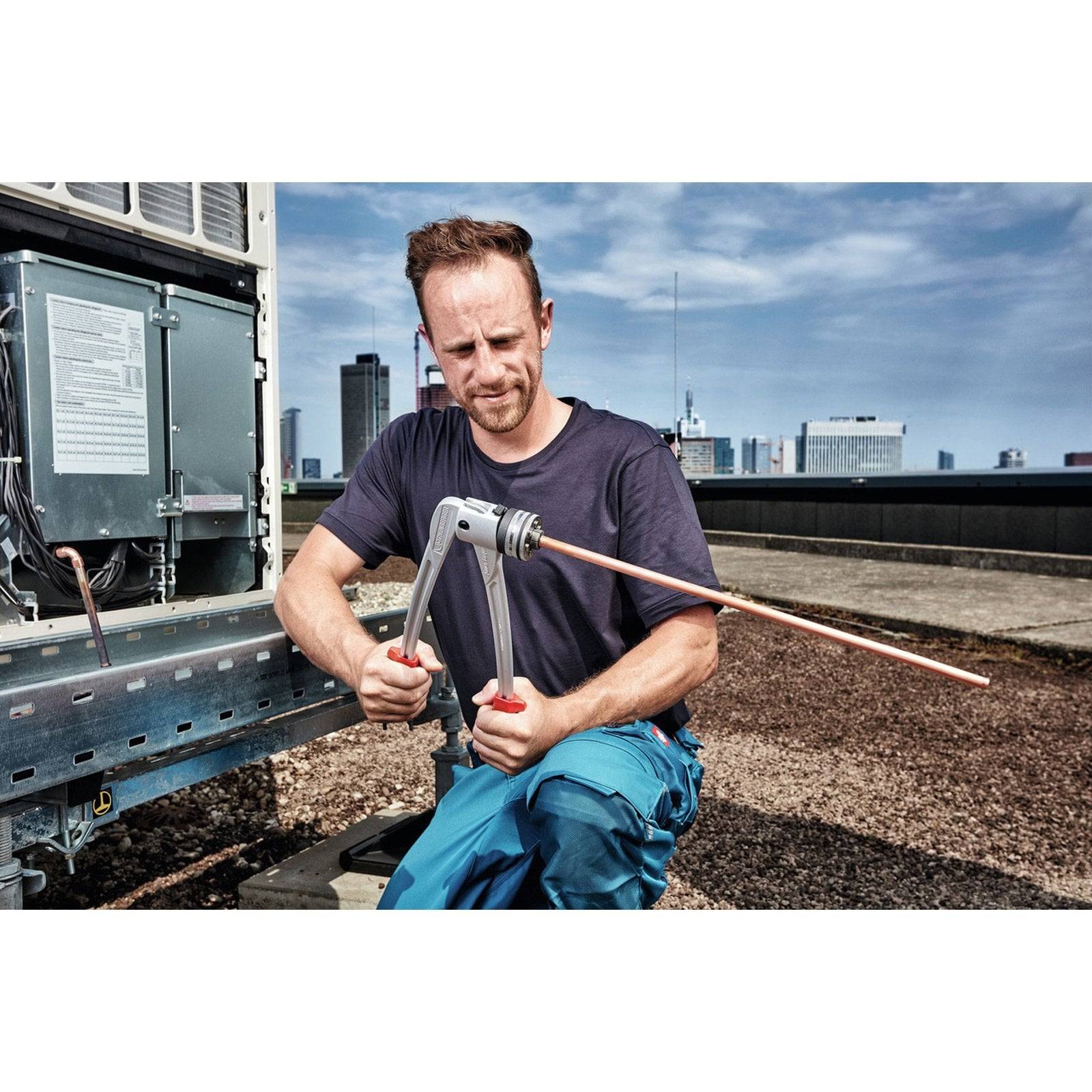 Eine Person biegt ein Kupferrohr auf einem Dach in der Nähe einer Klimaanlage, mit einer Stadtsilhouette im Hintergrund.