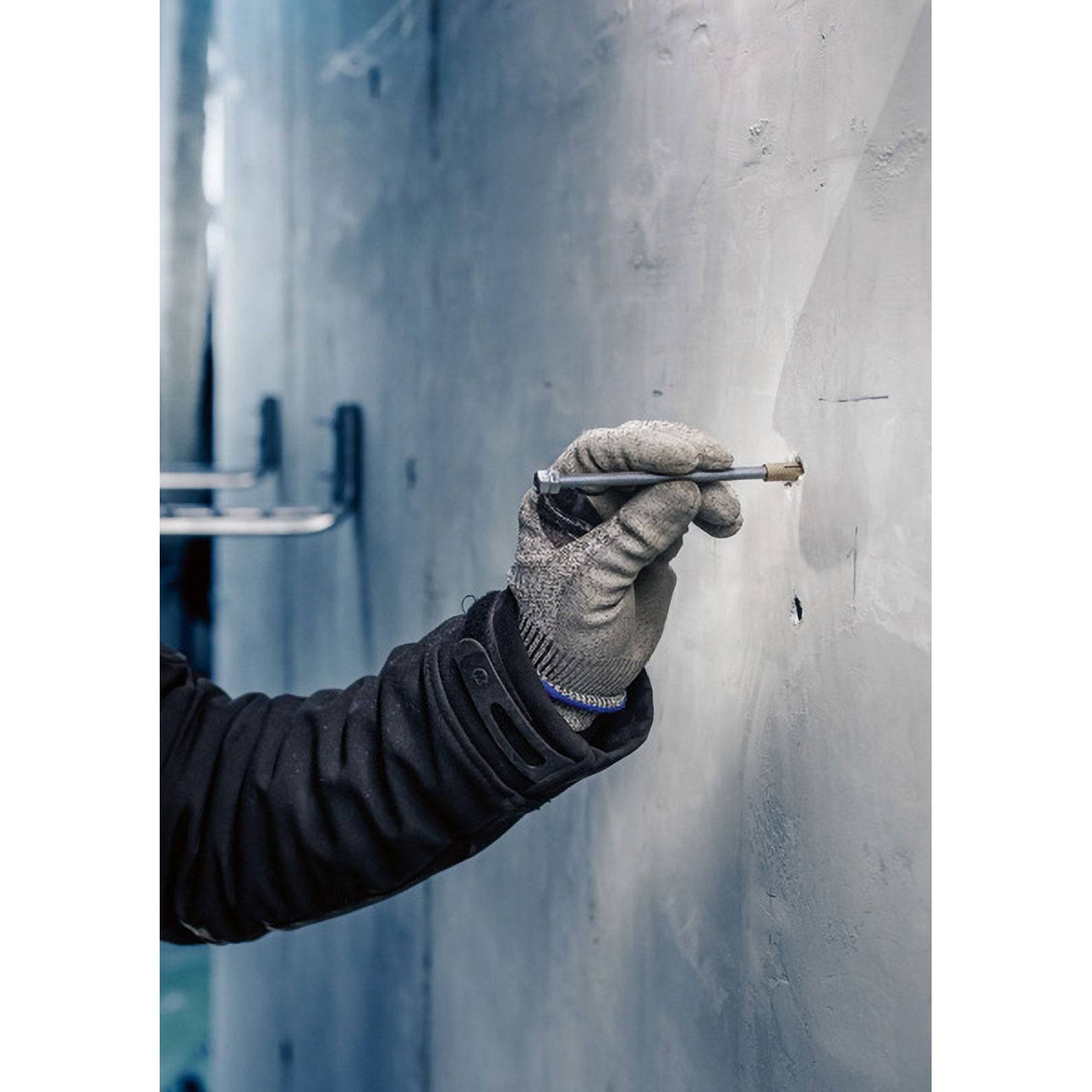 Eine Person trägt graue Handschuhe und eine dunkle Jacke und markiert eine Metalloberfläche mit einem Stift.