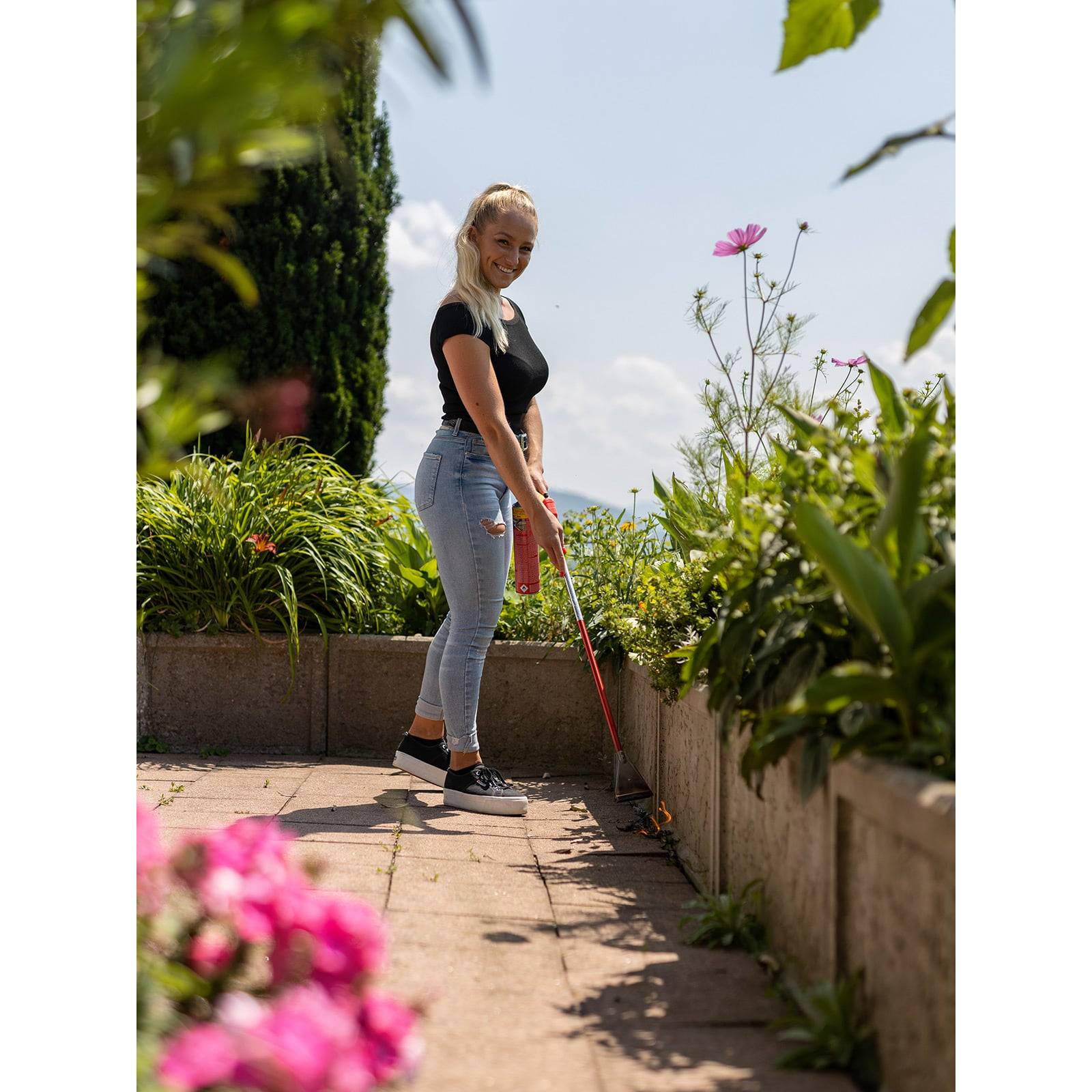 Eine Frau mit einem langen Werkzeug kehrt einen Gartenweg, der von üppigem Grün und Blumen umgeben ist, unter einem strahlend blauen Himmel.