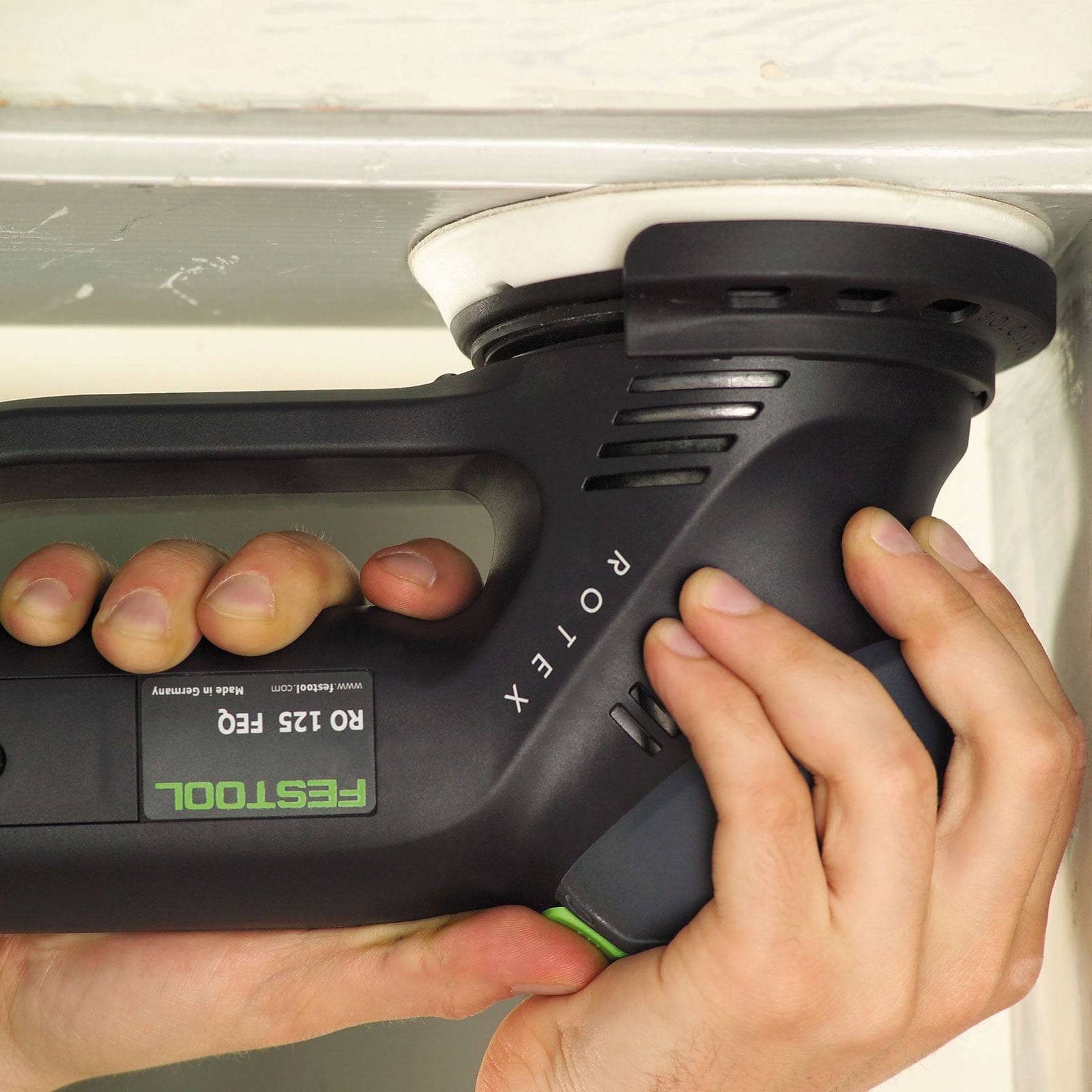 Eine Hand hält eine schwarze und grüne Exzenterschleifer mit der Bezeichnung ‚RO 125 FEQ Festool', der eine weiße Oberfläche poliert.