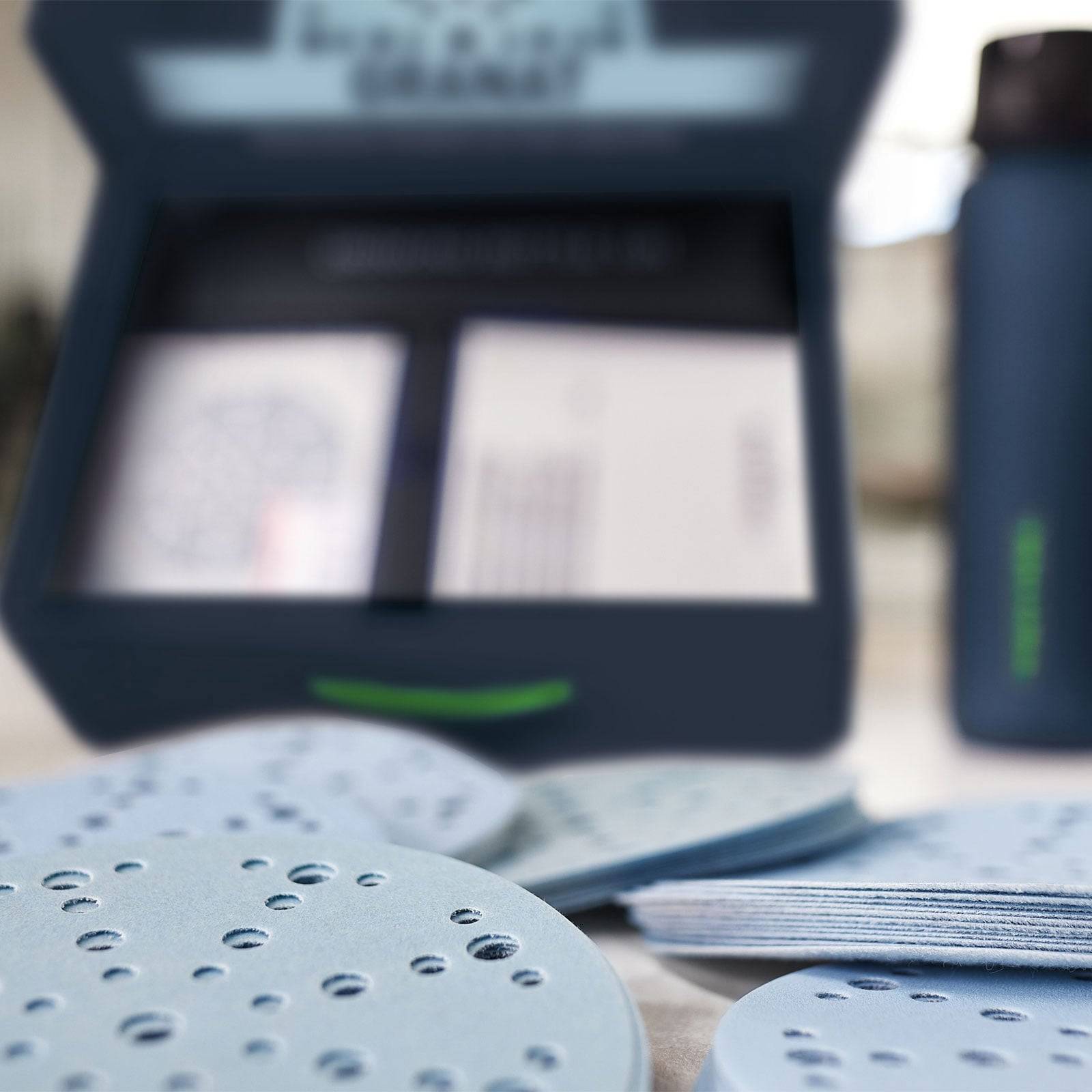 Eine Nahaufnahme von kreisförmigen Schleifpapieren und einer Schachtel in einer Werkstattsumgebung. Die Schleifpapiere haben mehrere Löcher, was möglicherweise auf eine spezifische Verwendung hindeutet.