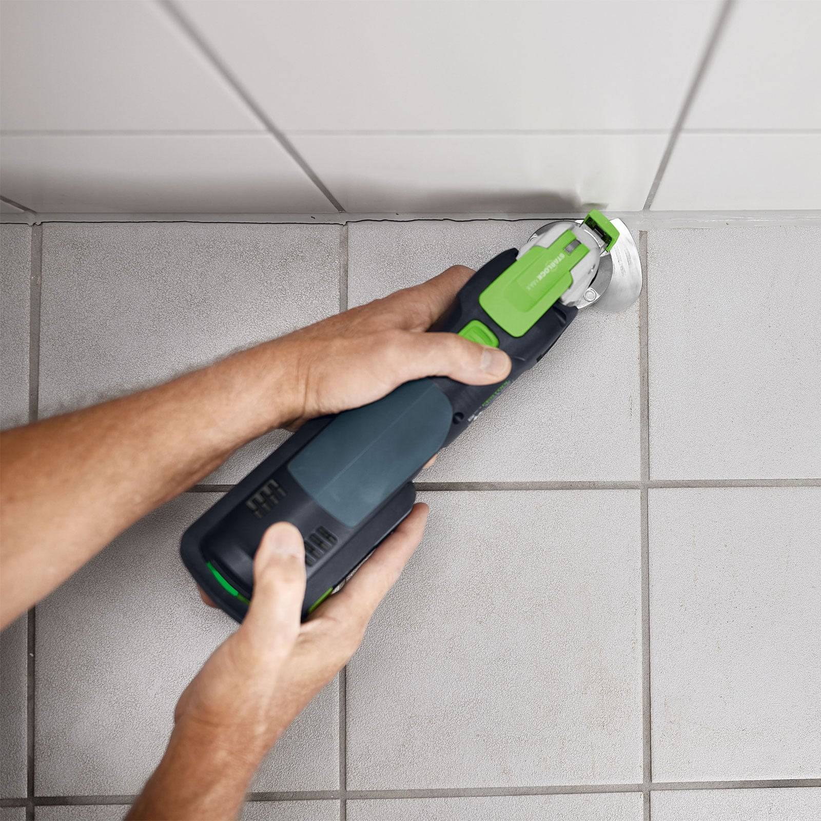 Eine Person hält ein Elektrowerkzeug und schleift den Mörtel zwischen Bodenfliesen an einer gefliesten Wand, was auf Hausrenovierung oder -reparatur hindeutet.