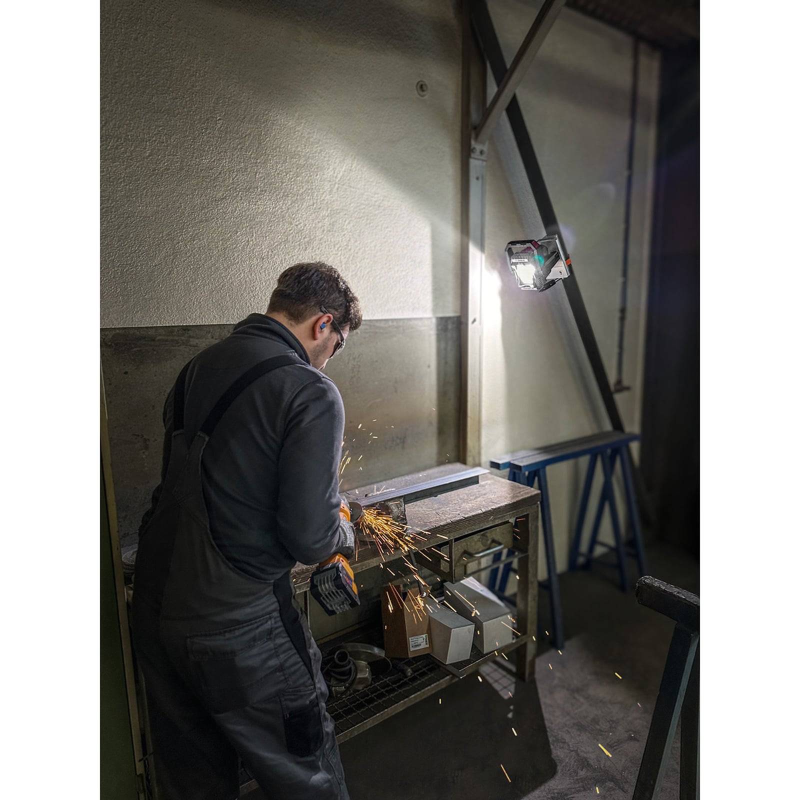 Eine Person in Arbeitskleidung verwendet ein Schleifwerkzeug an Metall und erzeugt dabei Funken. Die Umgebung scheint eine Werkstatt mit verschiedenen Werkzeugen und Ausrüstungen zu sein.