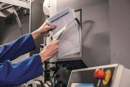 Eine Person in einer blauen Uniform legt einen Papierbogen auf eine Maschine oder inspiziert diesen, möglicherweise beschäftigt in einem Druck- oder Fertigungsprozess.