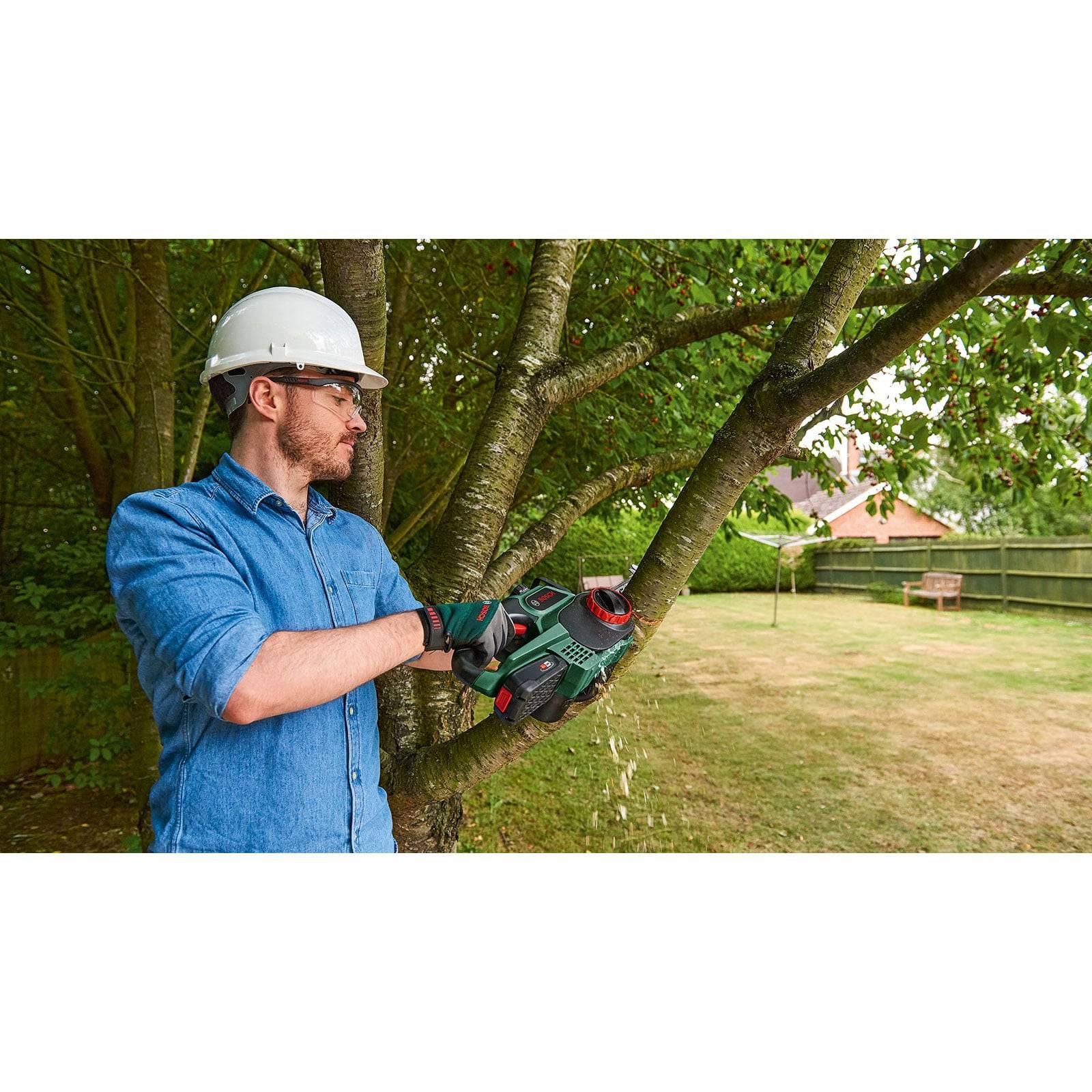 Eine Person mit Sicherheitsausrüstung verwendet eine Motorsäge, um Äste auf einem Baum in einem Hinterhof zu beschneiden, wobei der Schwerpunkt auf Rasenunterhalt und Sicherheit liegt.