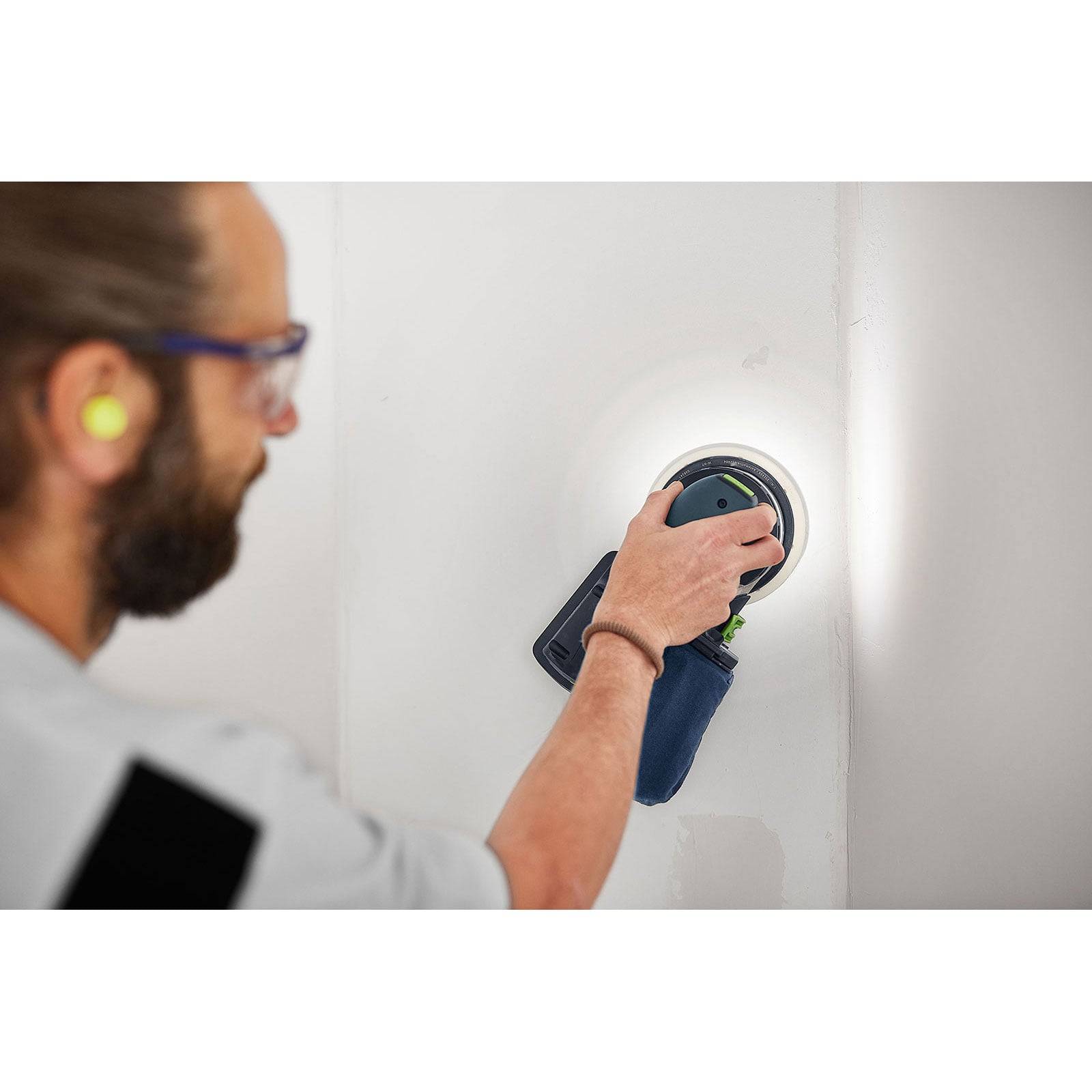 Person, die eine Wand mit einer elektrischen Schleifmaschine glättet und dabei Gehörschutz und eine Schutzbrille trägt, wobei er sich auf eine kreisförmige Bewegung konzentriert.