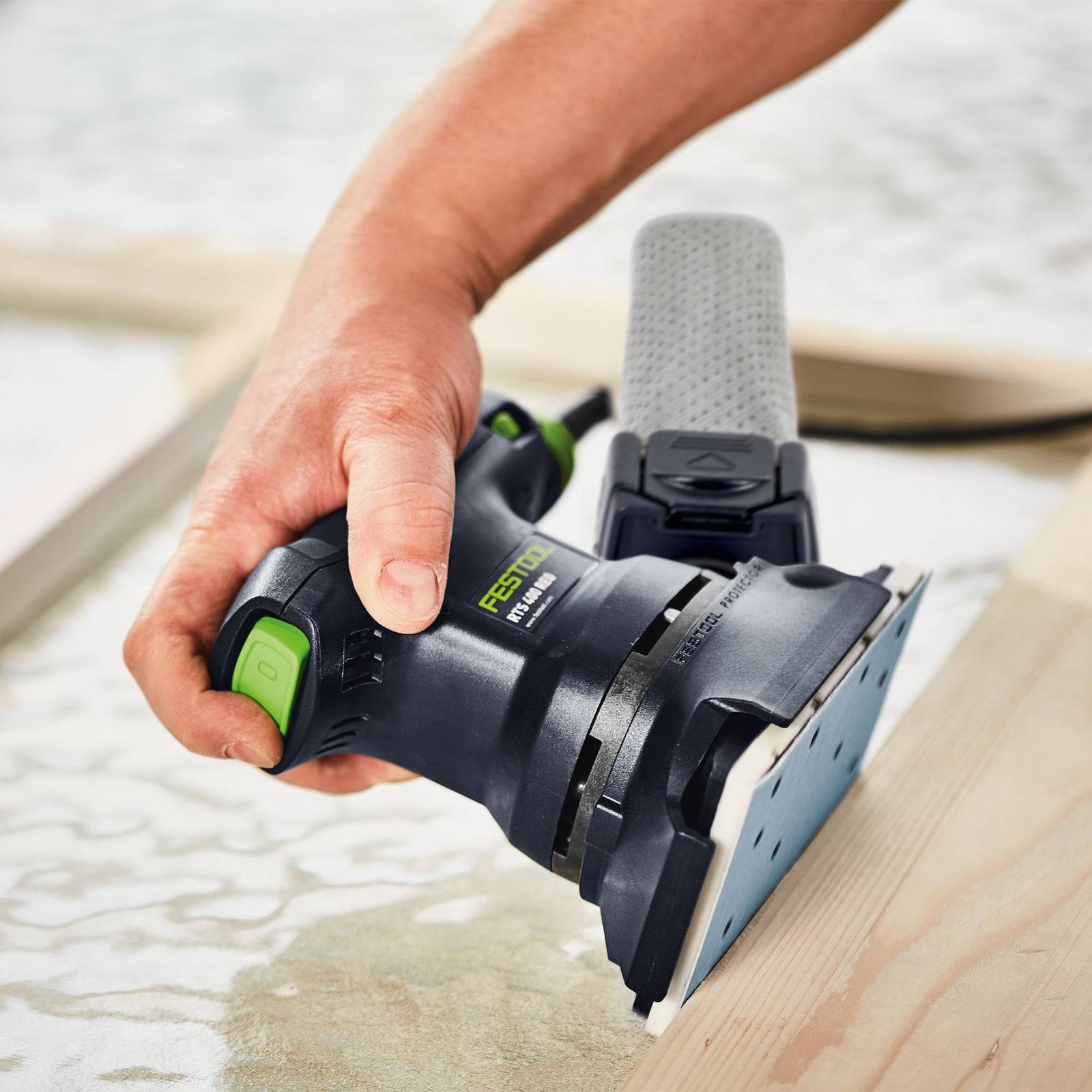Eine Person verwendet eine elektrische Schleifmaschine auf einem Holzbrett und bereitet die Oberfläche vor. Die Schleifmaschine ist schwarz und grün und verfügt über einen Staubauffangaufsatz.