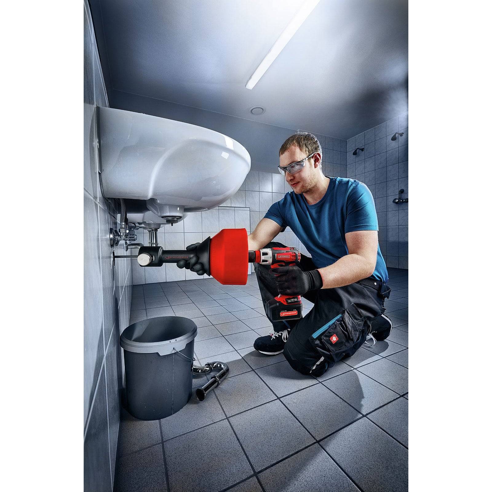 Eine Person mit Handschuhen benutzt ein Elektrowerkzeug, um Sanitärarbeiten unter einem Waschbecken in einem gefliesten Badezimmer durchzuführen. Ein Eimer steht auf dem Boden in der Nähe.