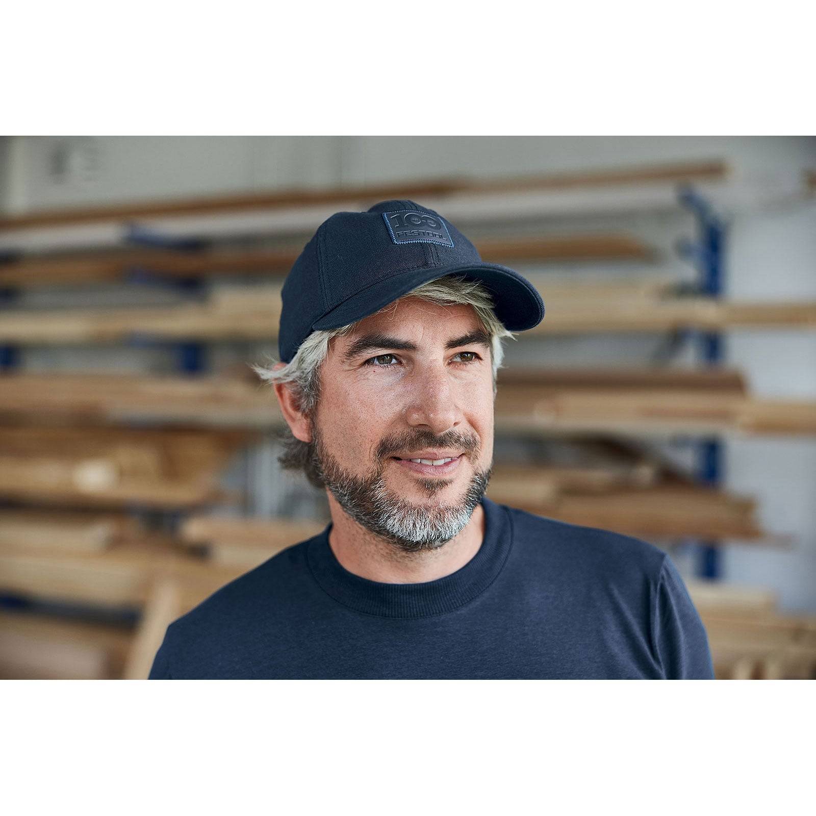 Ein Mann mit Bart, der eine dunkelblaue Mütze und ein dunkelblaues Hemd trägt, steht in einer Werkstatt mit Holzbrettern im Hintergrund.