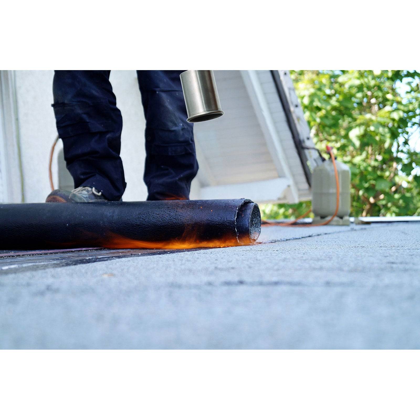 Ein Arbeiter verwendet einen Brenner, um Dachdeckmaterial auf einer flachen Oberfläche zu versiegeln, mit einer Leiter und Bäumen im Hintergrund.