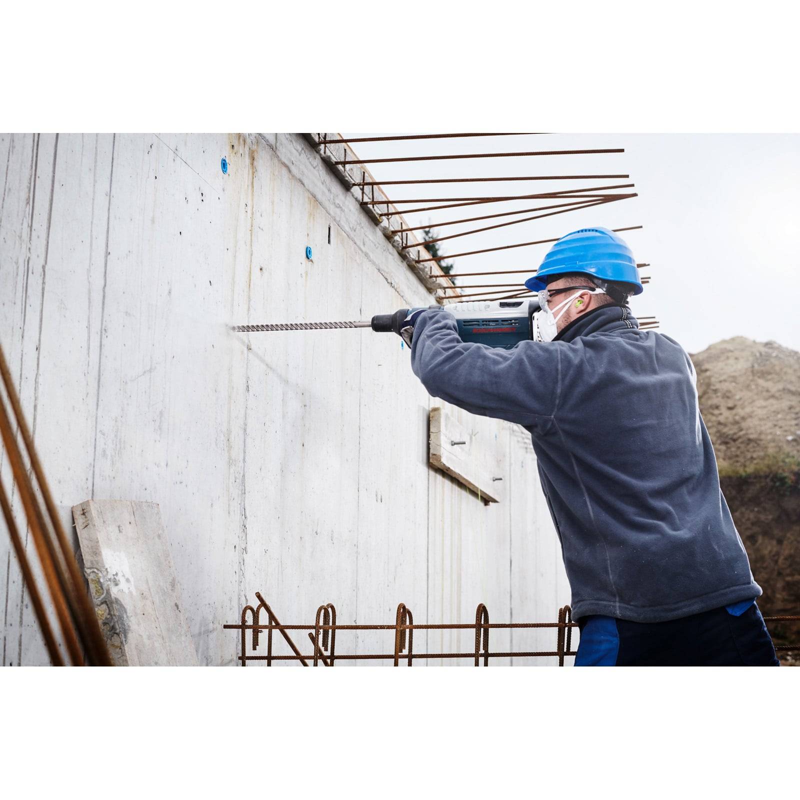 Ein Bauarbeiter in Schutzausrüstung bohrt in eine Betonwand, umgeben von freiliegendem Metallbewehrungsstahl, und unterstreicht damit industrielle Arbeit.