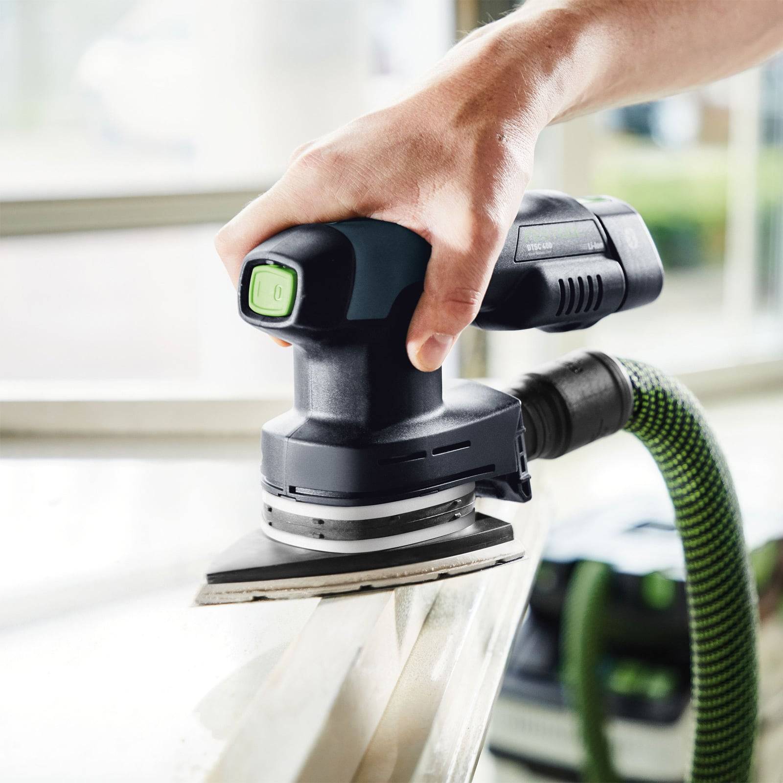 „Eine Hand, die einen schwarzen und grünen elektrischen Schleifer auf einer Holzoberfläche verwendet, mit einem grünen Schlauch für Staubabsaugung angeschlossen.