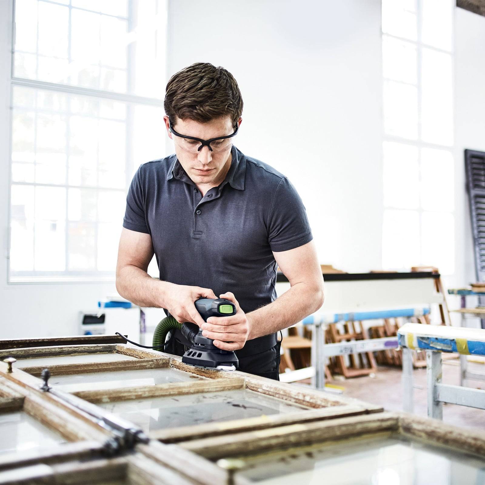Eine Person mit Sicherheitsbrille bedient eine Maschine an einem Glasstück in einer Werkstatt, was Präzision und Konzentration im Handwerk andeutet.
