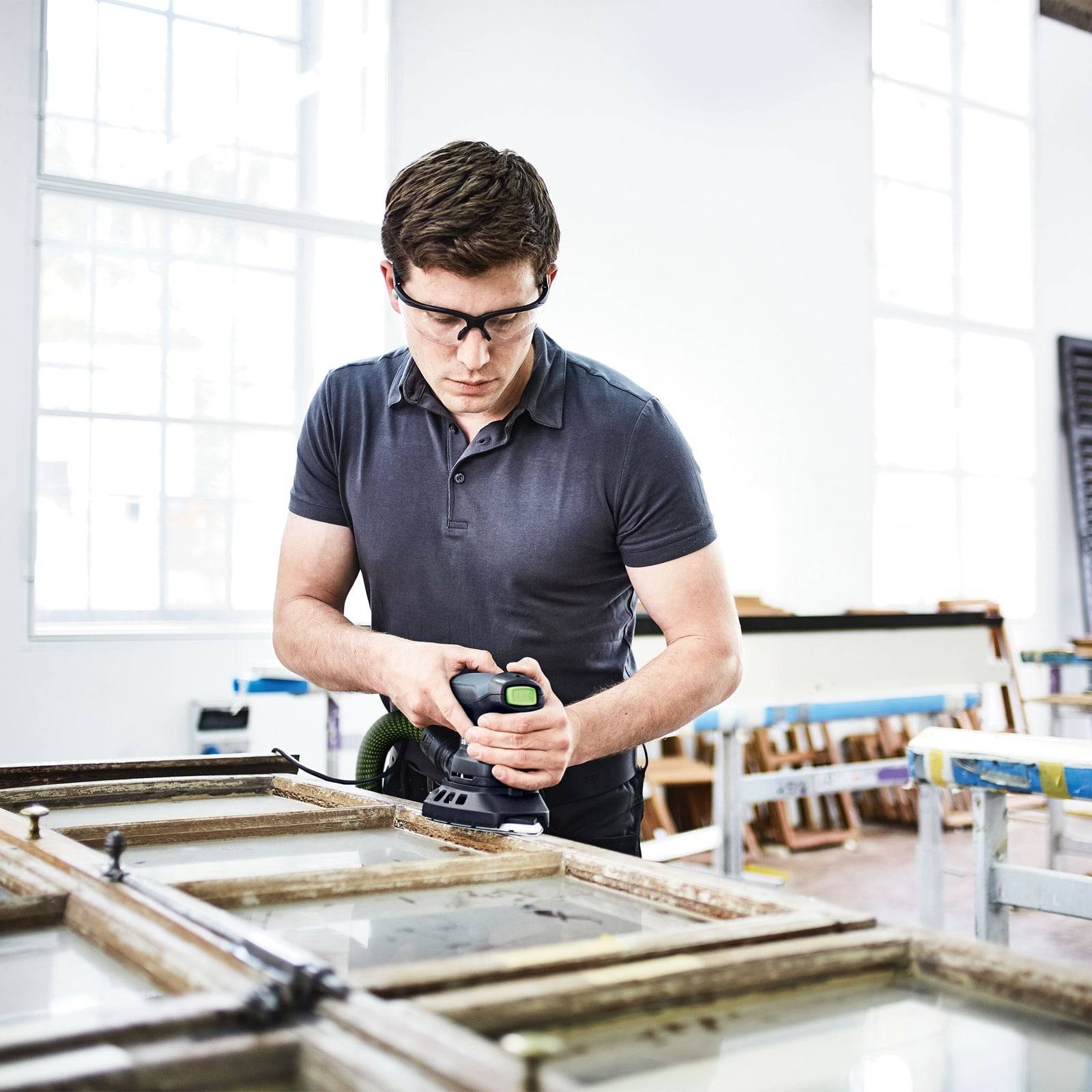 Eine Person mit Sicherheitsschutzbrille benutzt ein Elektrowerkzeug, um an einer großen Fensterscheibe in einer hellen Werkstatt zu arbeiten.