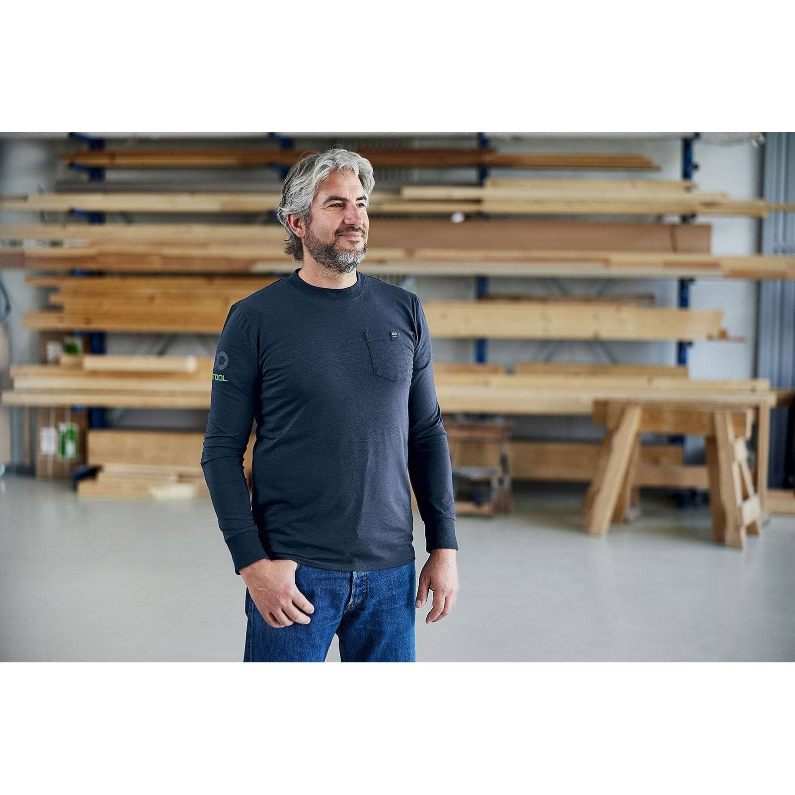 Eine Person mit grauen Haaren und einem Bart steht in einer Werkstatt und trägt ein langärmliges Hemd und Jeans, umgeben von Holzbalken.