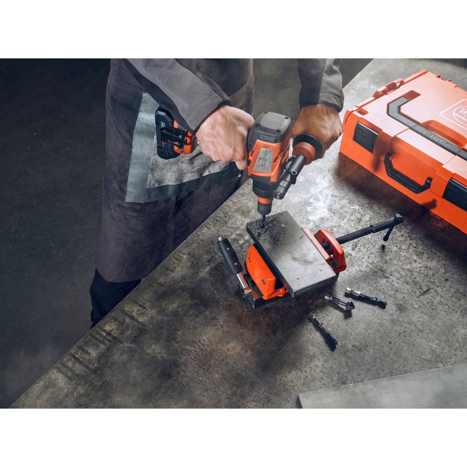 Eine Person benutzt einen orangefarbenen Akkuschrauber auf einem Metallobjekt, umgeben von Bohrern und einer orangefarbenen Werkzeugbox auf einer Werkbank.