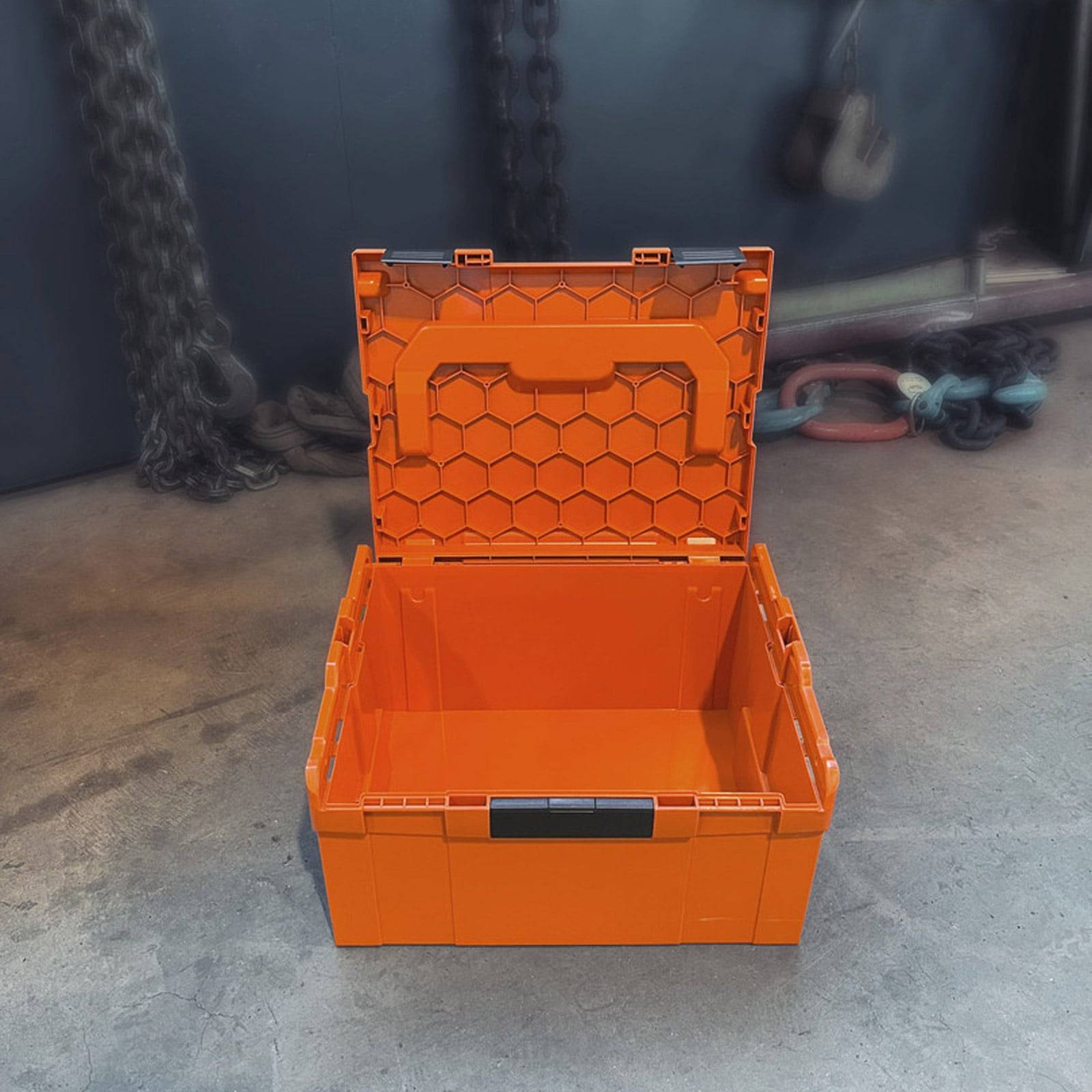 Eine orangefarbene Aufbewahrungsbox mit einem Wabenmuster-Deckel ist geöffnet und steht auf einem Betonboden. Industrieketten und Haken sind im Hintergrund sichtbar.