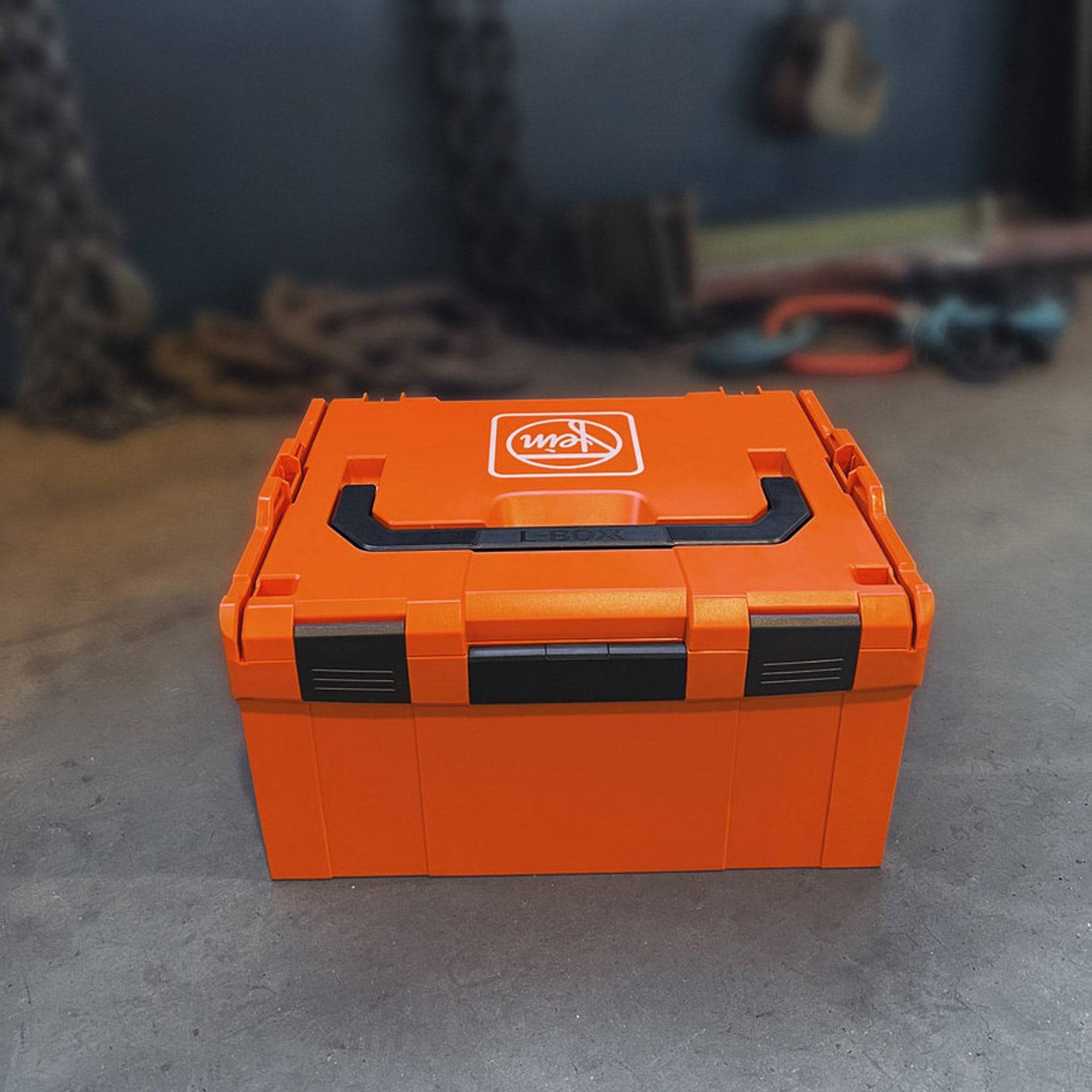 Orangefarbene Werkzeugkiste auf einem Betonboden, teilweise geöffnet mit einem sichtbaren Logo auf der Oberseite. Werkzeuge und Ketten sind im Hintergrund verstreut.