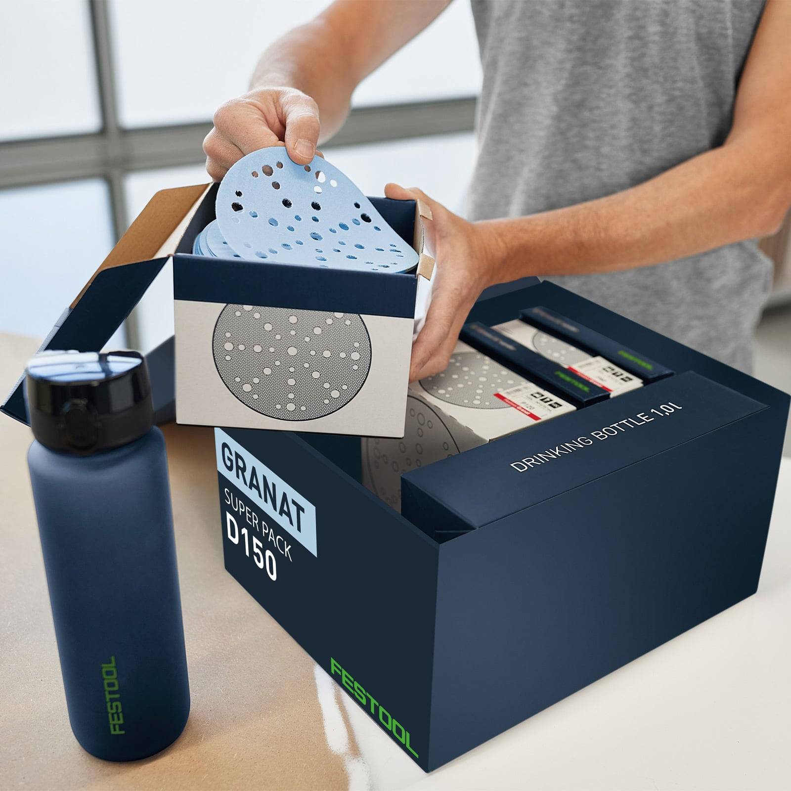 Eine Person legt Schleifteller in eine Festool-Box 'Granat Super Pack' mit 'TRINKFLASCHEN UND T-SHIRT' auf der Seite, die sichtbar sind.