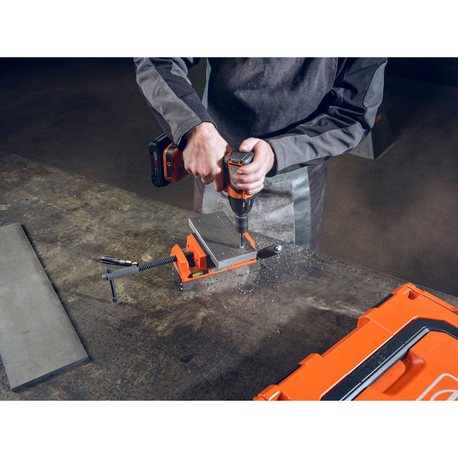 Eine Person in Arbeitskleidung bedient einen Akkuschrauber an einem Metallstück, das in einem Schraubstock auf einer Werkbank befestigt ist, umgeben von verschiedenen Werkzeugen.