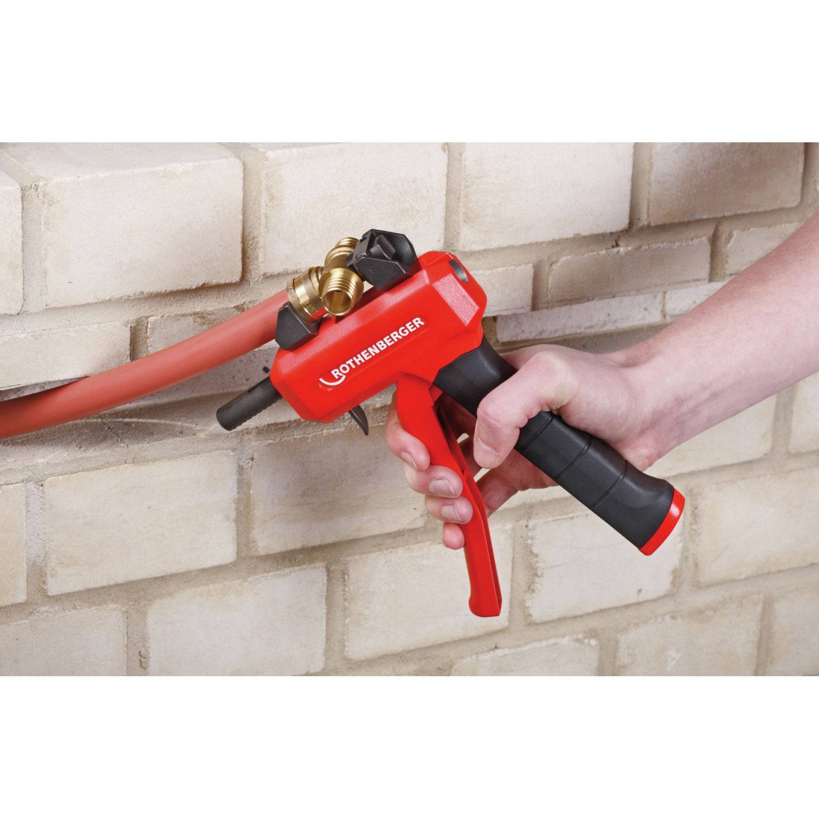 Eine Person verwendet ein rotes Werkzeug, um eine Armatur an einem roten Rohr zu befestigen, das sich von einer Ziegelmauer erstreckt. Das Werkzeug ist mit „ROTHENBERGER