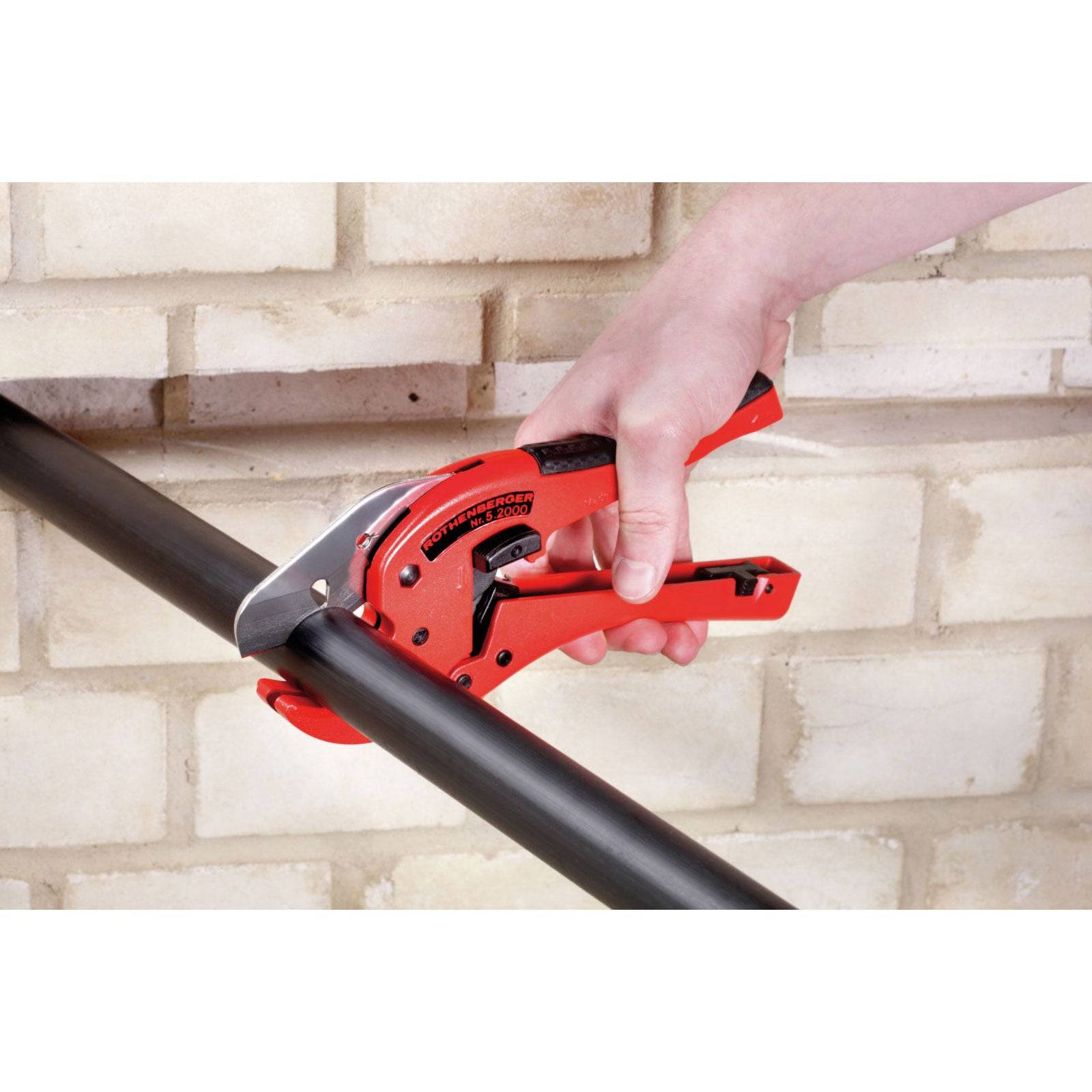 Eine Hand verwendet einen roten Rohrabschneider, um ein schwarzes Rohr vor einer Ziegelwand zu durchtrennen.