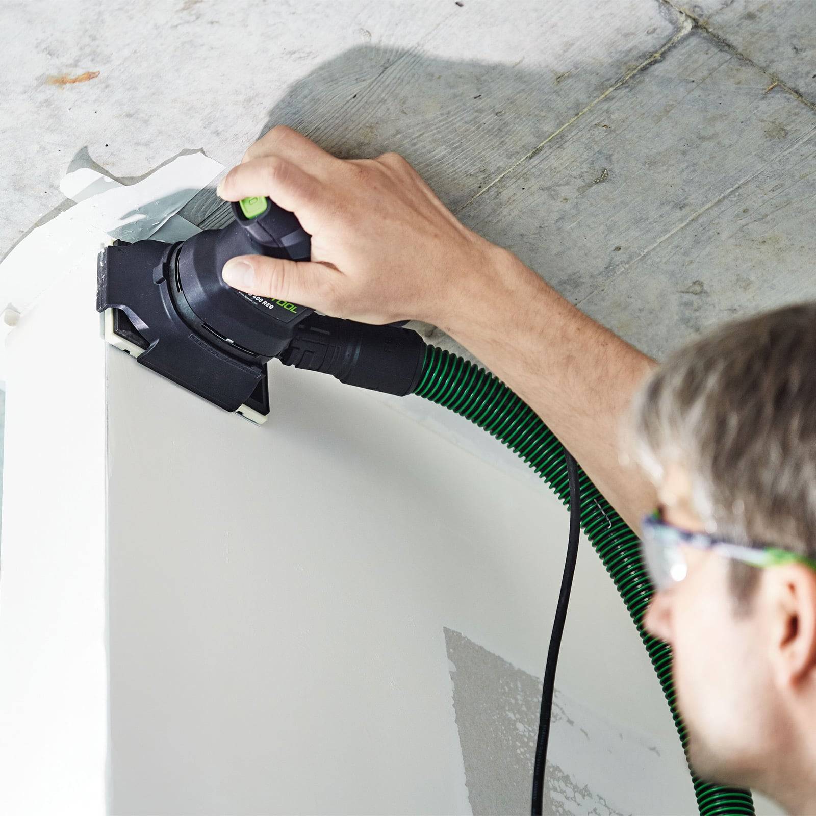 Eine Person, die ein Elektrowerkzeug verwendet, um eine Wandecke zu schleifen, während Staub durch einen angeschlossenen Saugschlauch aufgesammelt wird.