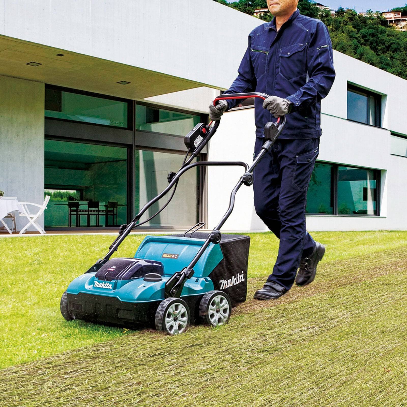 Eine Person in blauer Arbeitskleidung mäht mit einem elektrischen Rasenmäher einen Rasenplatz vor einem modernen weißen Haus mit großen Fenstern.