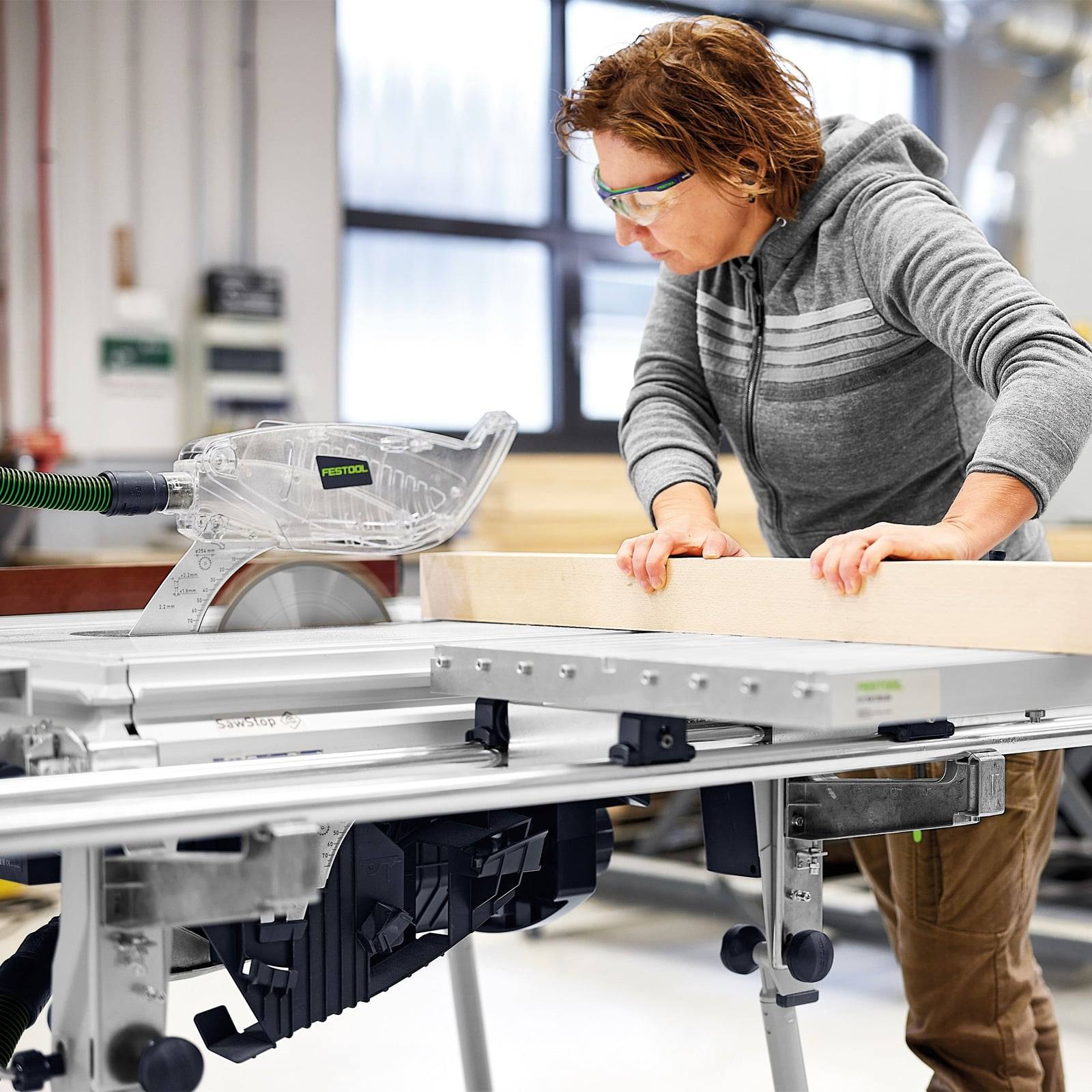 Eine Person in einem grauen Hoodie bedient eine Tischkreissäge und schneidet ein Stück Holz in einer Werkstatt. Der Fokus liegt auf Sicherheit und Präzision.