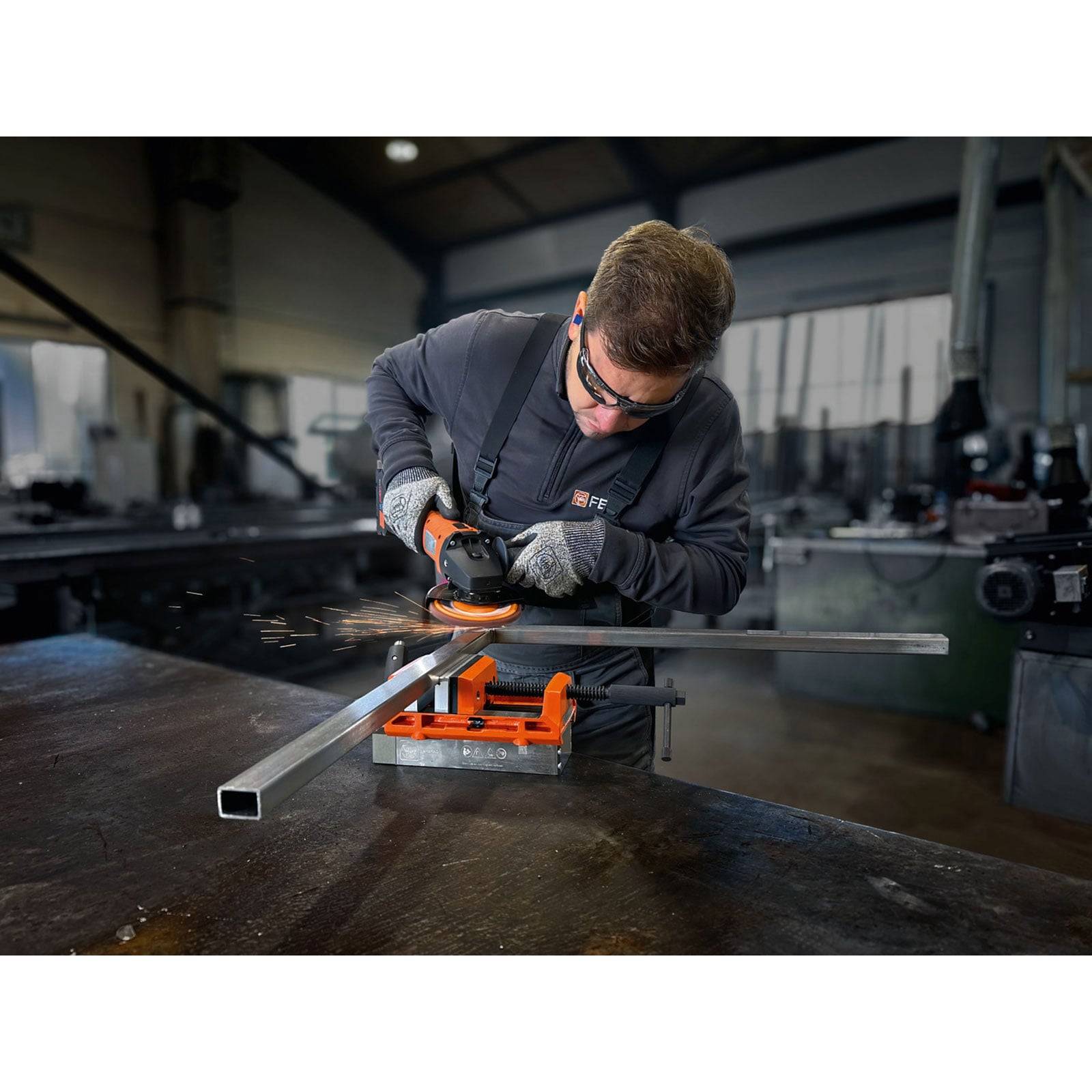 Ein Mann in Sicherheitsausrüstung verwendet eine Winkelschleifer an einem Metallstab in einer Industriewerkstatt. Funken sprühen, während er an einem Metalltisch arbeitet.