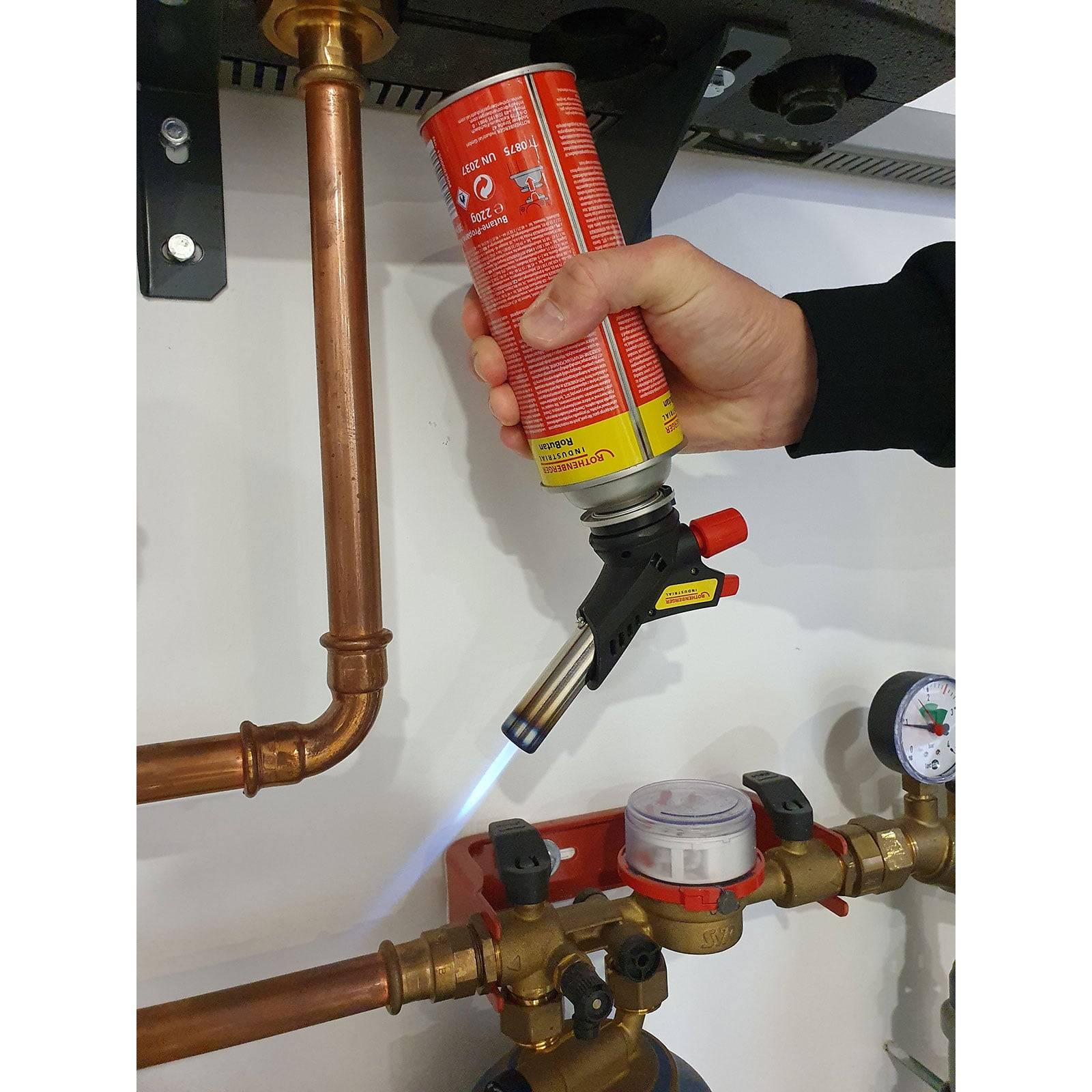 Eine Hand verwendet eine an einer Gaskartusche befestigte Fackel und erhitzt Kupferrohre in einer Klempnerinstallation. Manometer und Ventile sind sichtbar.