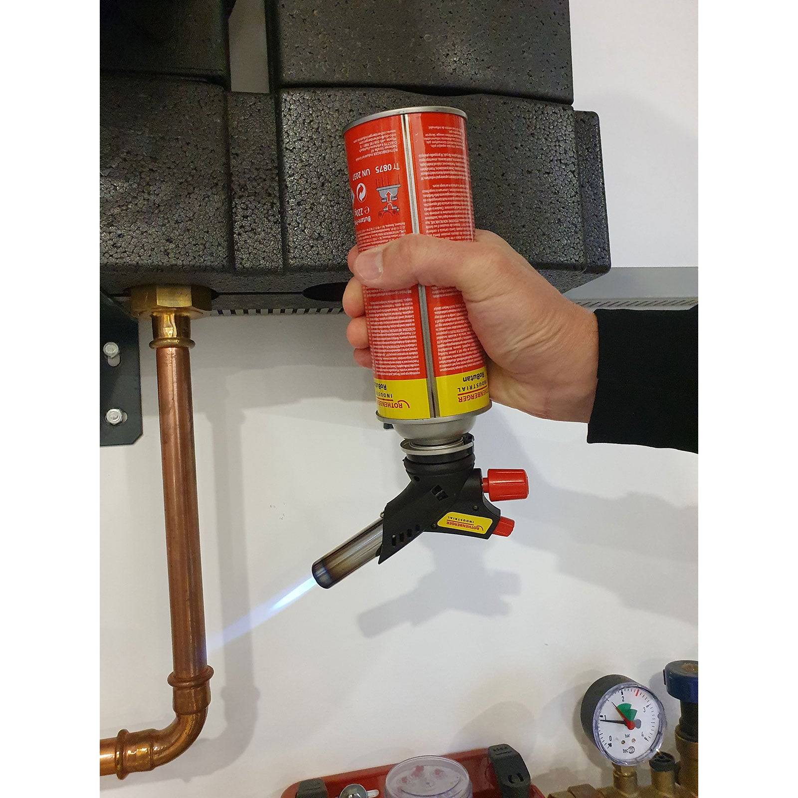 Eine Hand verwendet einen Propanbrenner, um ein Kupferrohr zu löten und emittiert dabei eine blaue Flamme. In der Nähe sind ein Druckmesser und schwarze Isolierung sichtbar.