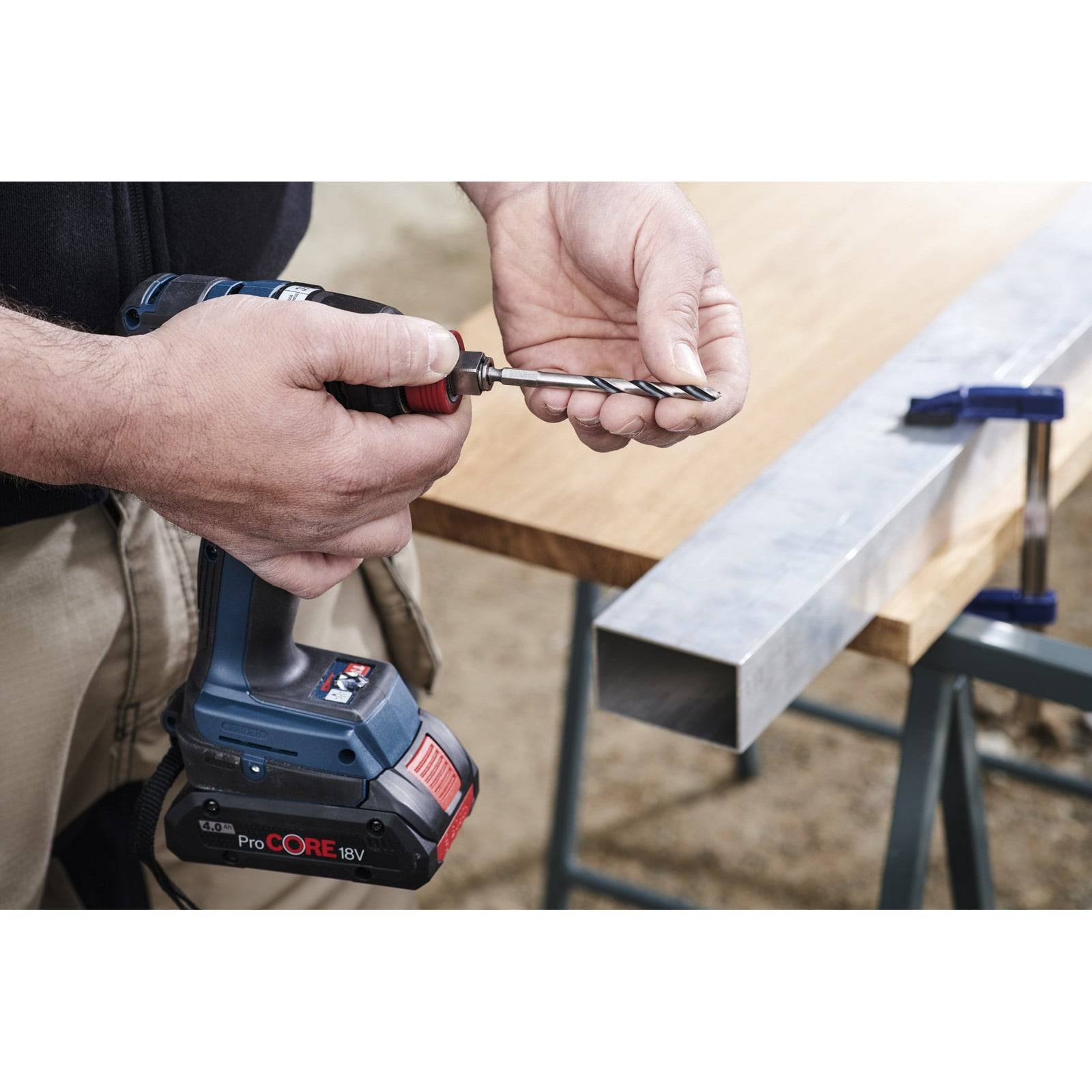 Eine Person, die einen kabellosen Akkuschrauber mit einem Metallbohrer verwendet, der daran befestigt ist und sich anschickt, in ein Metallwerkstück zu bohren, das auf einer Werkbank eingespannt ist.