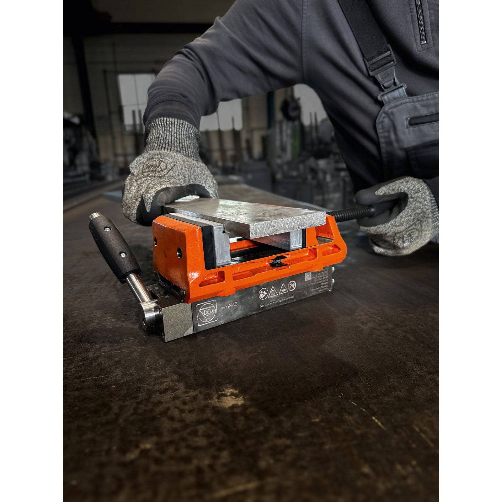 Eine Person in Handschuhen und Overall bedient einen schweren Hebemagneten auf einer Metalloberfläche und deutet damit auf eine industrielle oder Werkstattnutzung hin.