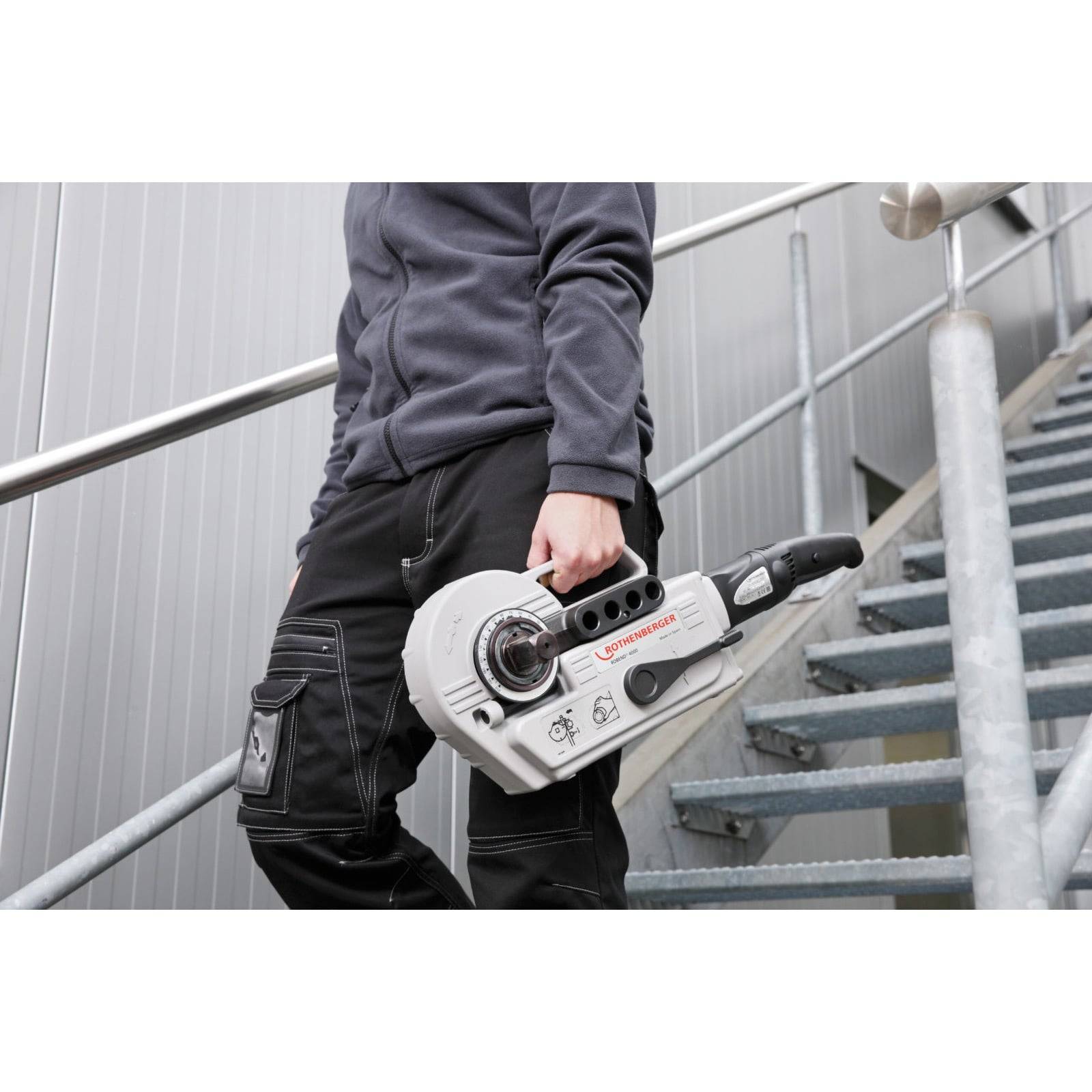 Eine Person, die auf einer Treppe steht und ein industrielles Werkzeug hält, trägt eine graue Jacke und schwarze Hosen. Das Werkzeug ist mit dem Markennamen 'RIDGID' gekennzeichnet.