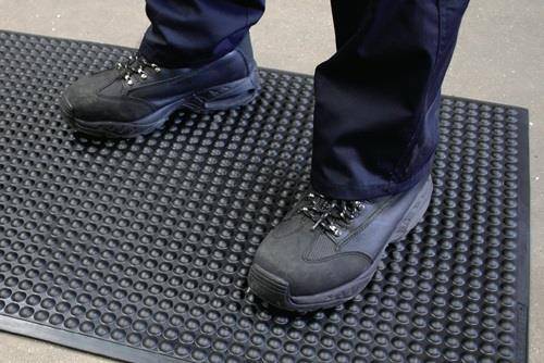 Person, die auf einer strukturierten Anti-Ermüdungsmatte steht und schwarze Schuhe und dunkle Hose trägt.