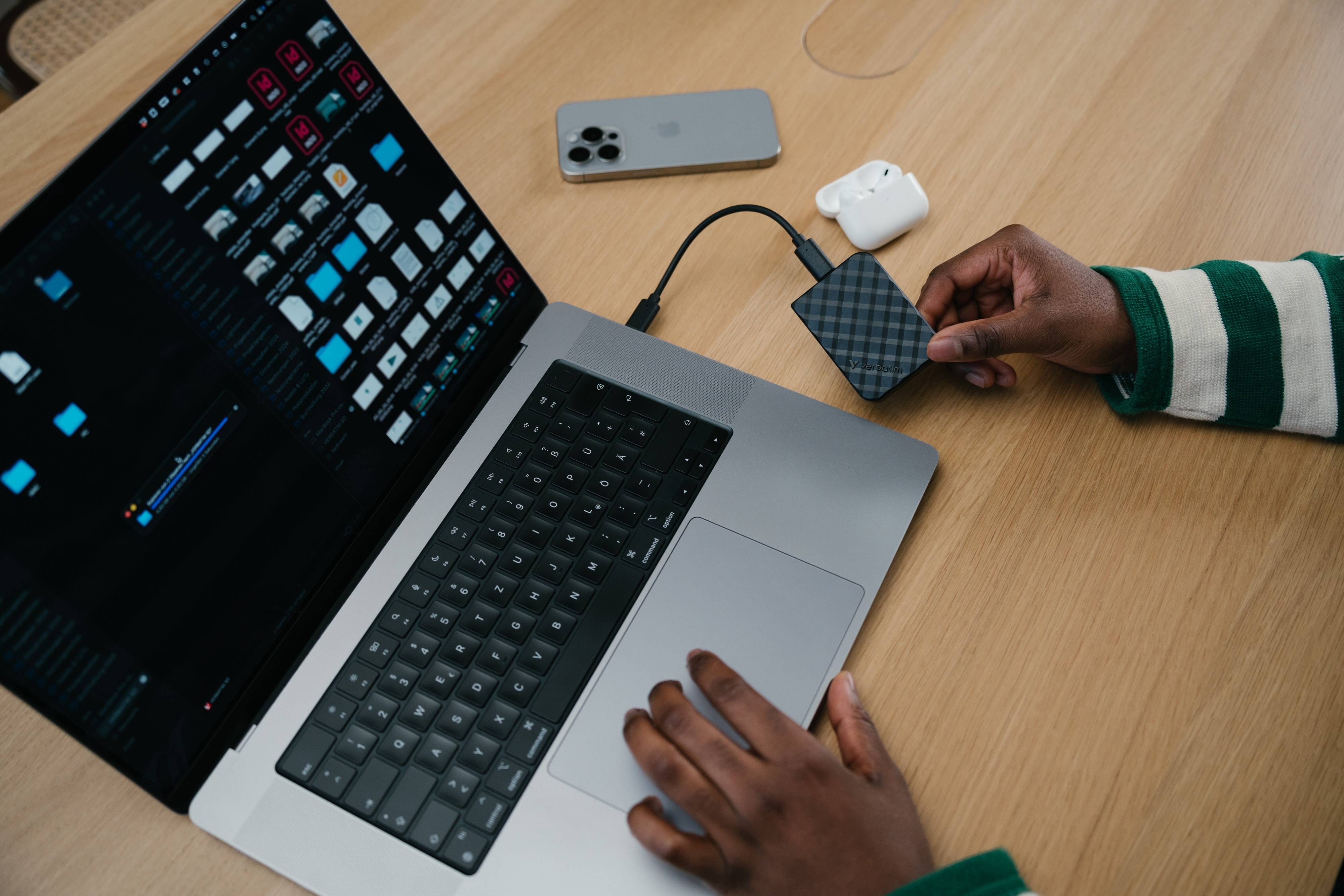 Eine Person verbindet ein kleines tragbares Laufwerk mit einem Laptop über ein USB-Kabel auf einem Schreibtisch, mit einem Smartphone und Kopfhörern in der Nähe.