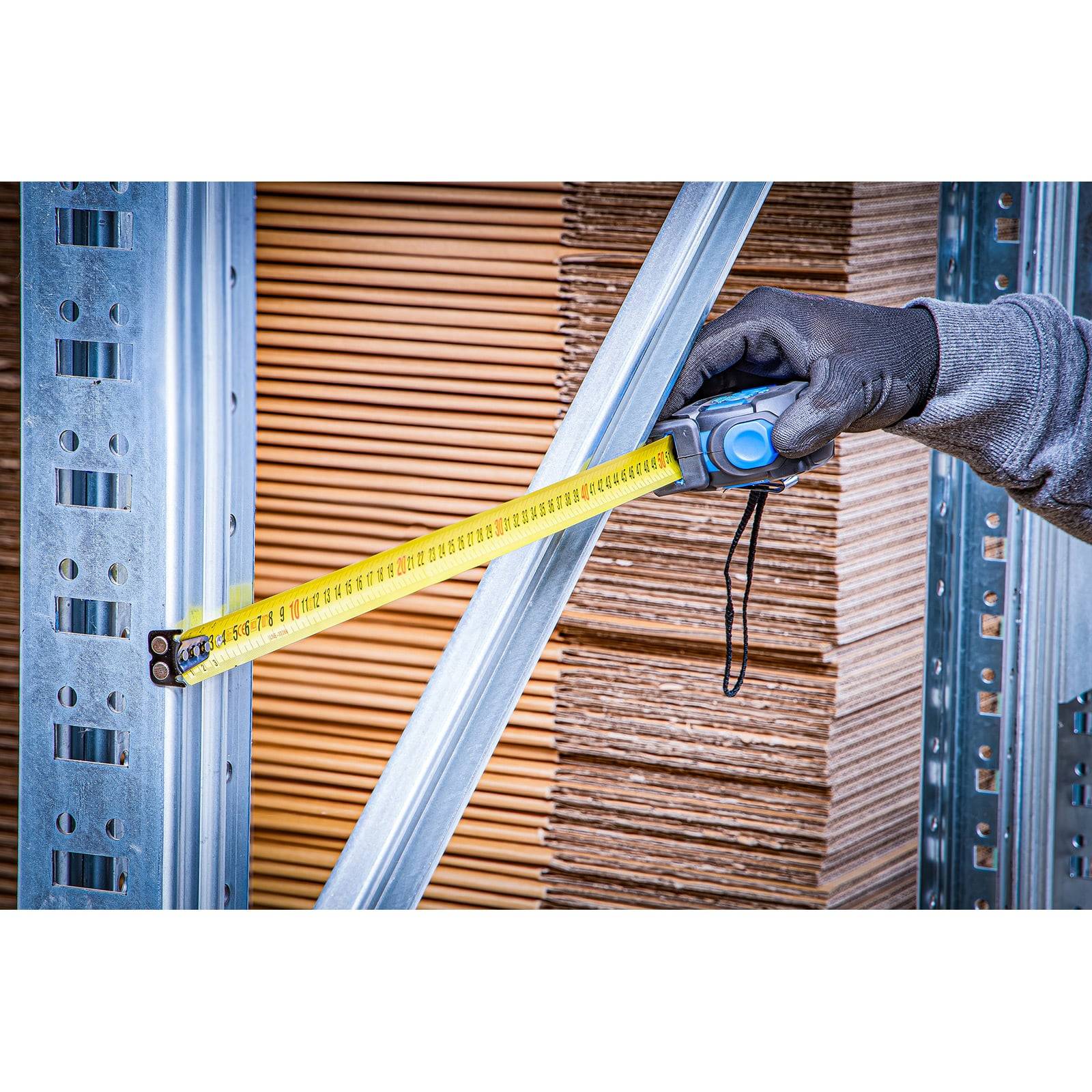 Eine Person misst Metallregale mit einem gelben Maßband in einem Lagerraum, der mit Stapeln von Kartons gefüllt ist.