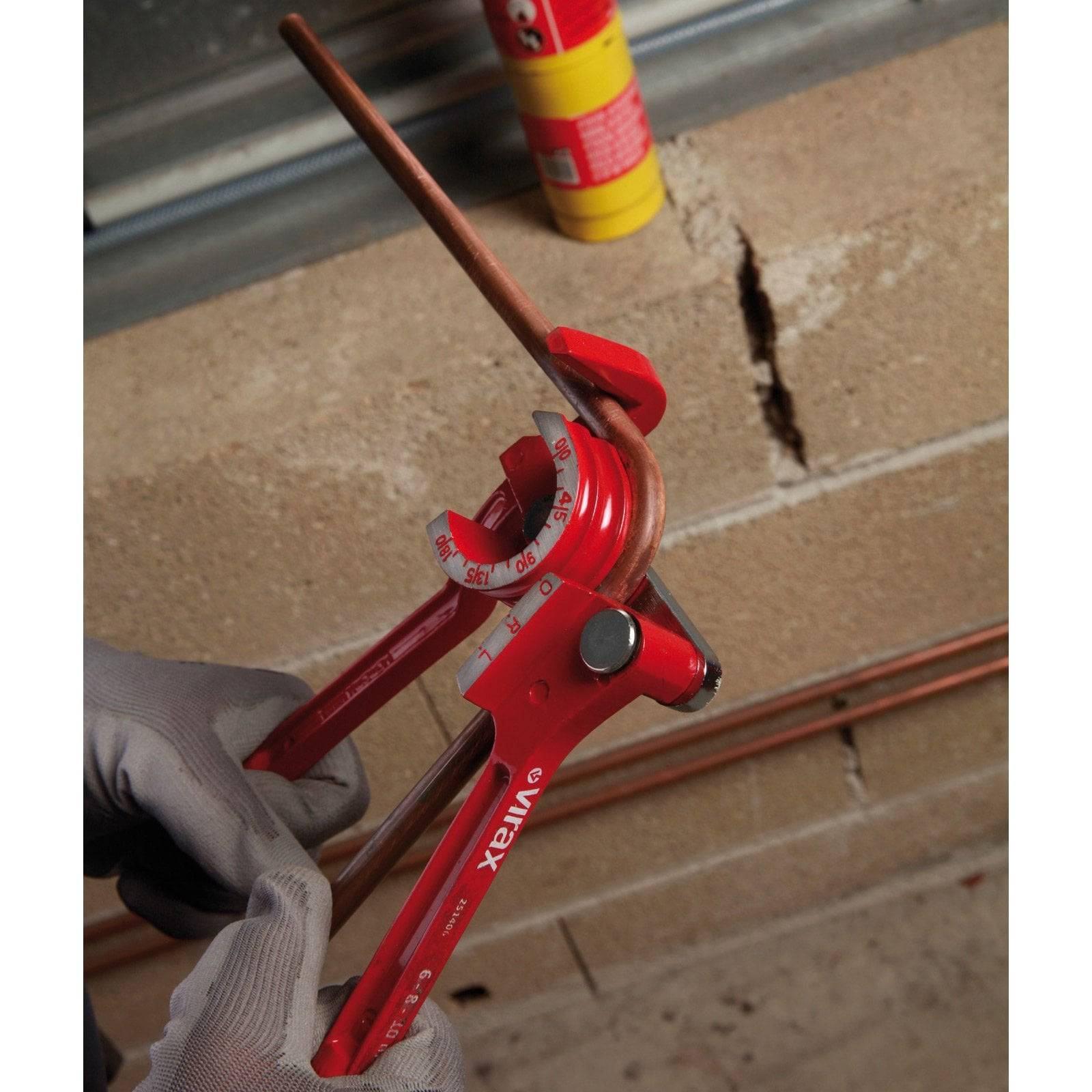 Eine Person biegt ein Kupferrohr mit einem roten manuellen Rohrbiegewerkzeug vor einer Ziegelwand.