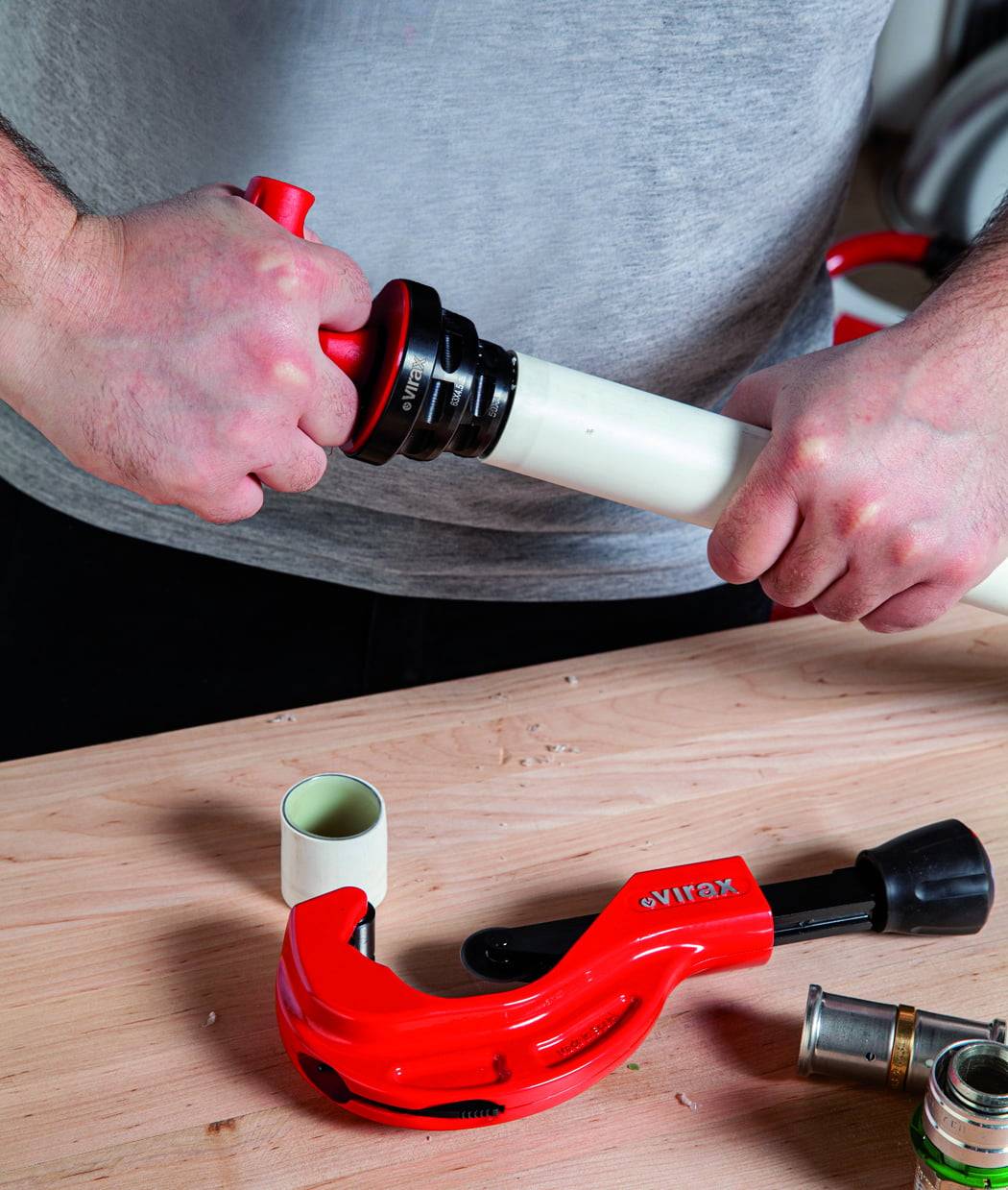 Zwei Hände, die ein weißes Rohr mit einem roten Werkzeug auf einer Holzoberfläche zusammenbauen, was auf Rohrreparatur oder sanitäre Arbeiten hinweist.