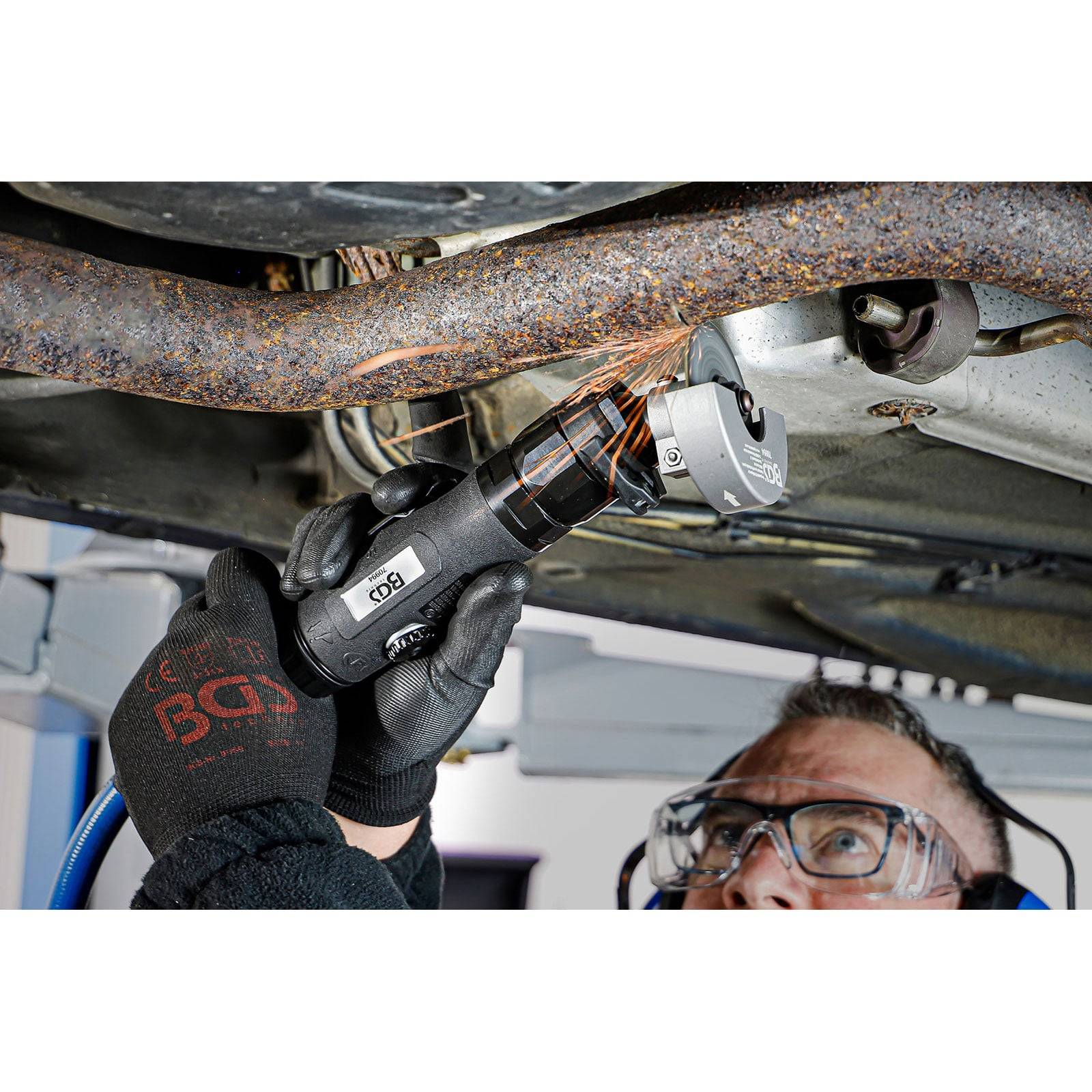 Eine Person mit Schutzbrille und Handschuhen verwendet ein Elektrowerkzeug, um ein rostiges Metallrohr unter einem Fahrzeug zu durchtrennen.