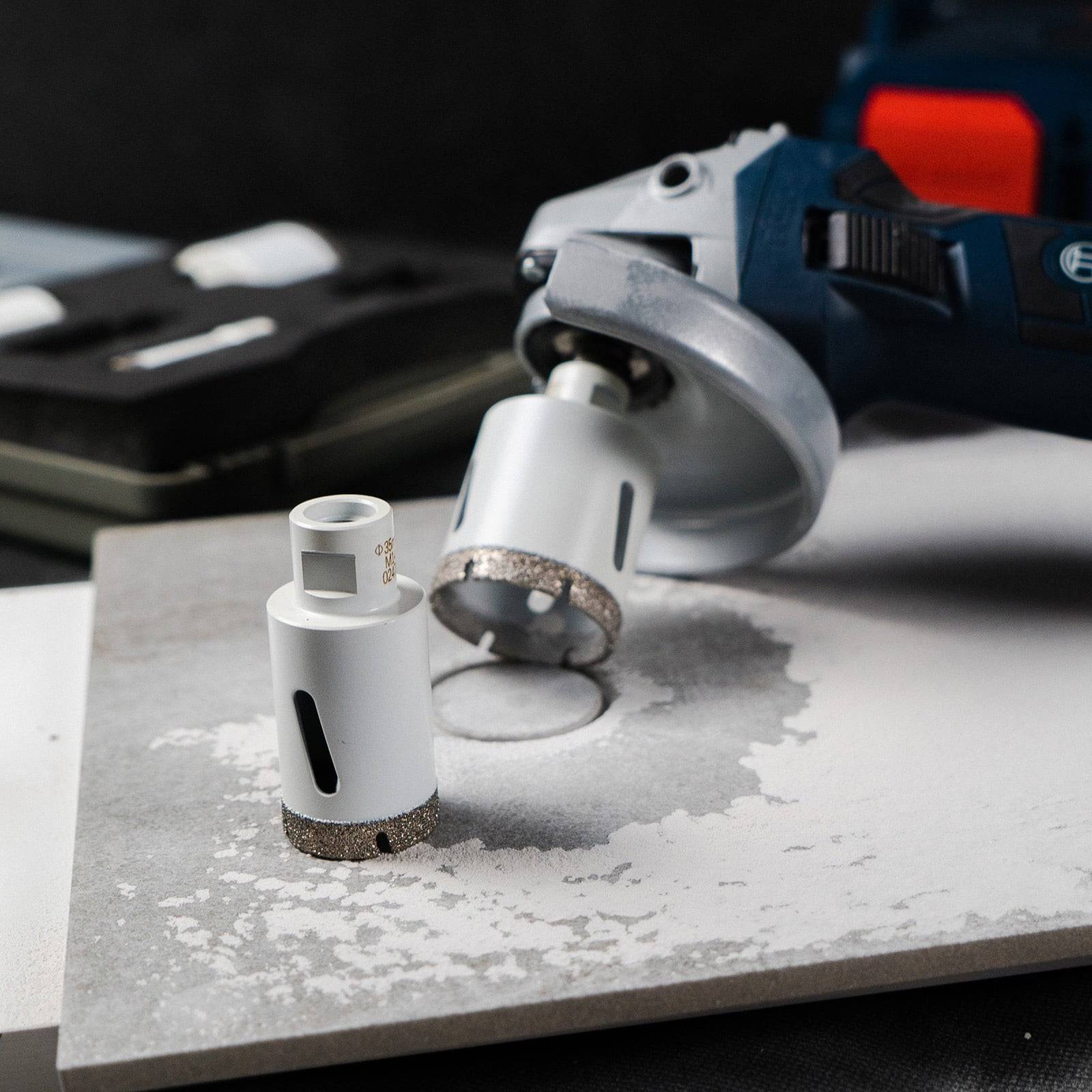 Ein elektrischer Schleifer mit einer Diamantlochsäge ist auf einer grauen Fliese abgelegt, umgeben von Staub und einer weiteren Lochsäge in der Nähe.