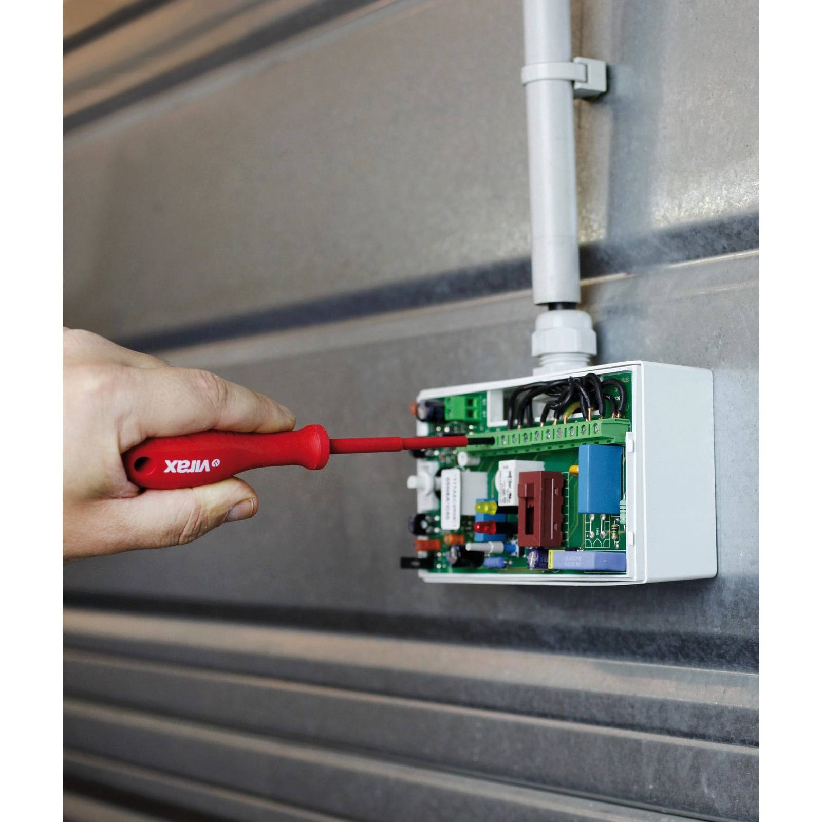 Eine Person verwendet einen roten Schraubendreher, um Komponenten in einem offenen Schaltkasten anzupassen, der an einer Metallwand montiert ist.