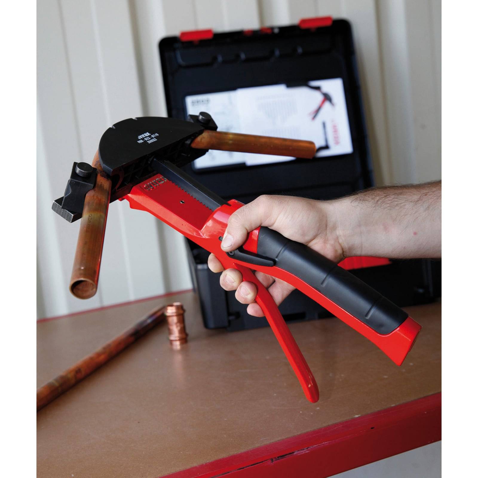 Eine Person hält einen roten und schwarzen Rohrbiegeapparat und demonstriert dessen Verwendung an einem Kupferrohr. Ein schwarzer Werkzeugkoffer befindet sich im Hintergrund.