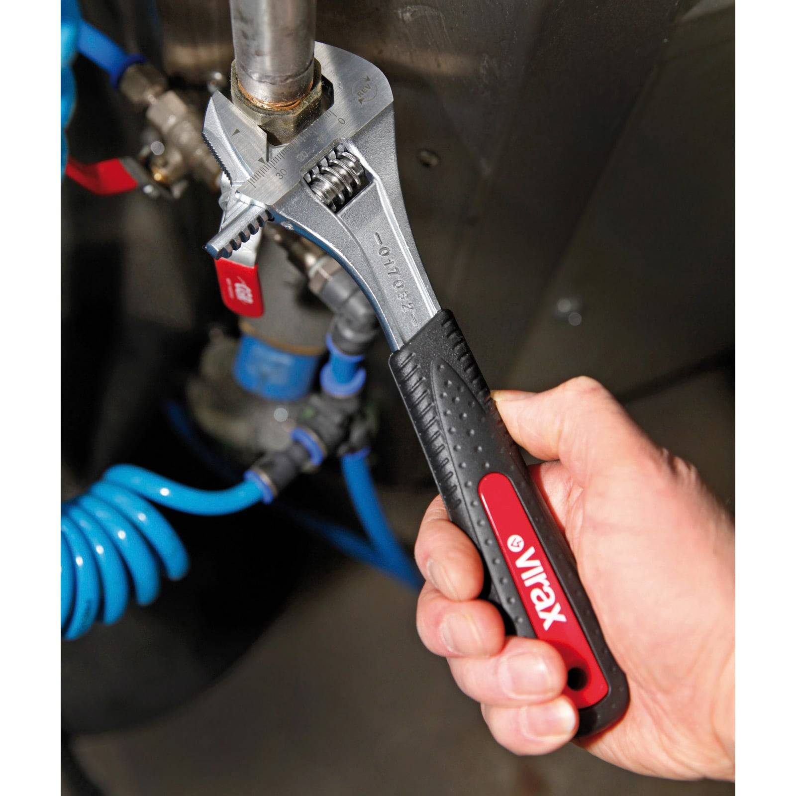 Eine Hand benutzt einen verstellbaren Schraubenschlüssel in Rot und Schwarz, um eine Rohranschlussverbindung zu befestigen, wobei im Hintergrund blaue Rohrleitungen sichtbar sind.