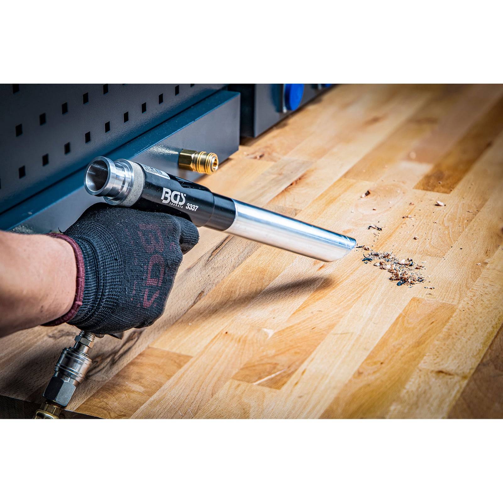 Eine Person verwendet einen schwarzen und silbernen pneumatischen Staubsauger an einem Holzarbeitstisch, um kleine Trümmer zu reinigen und trägt dabei einen schwarzen Handschuh zur Sicherheit.